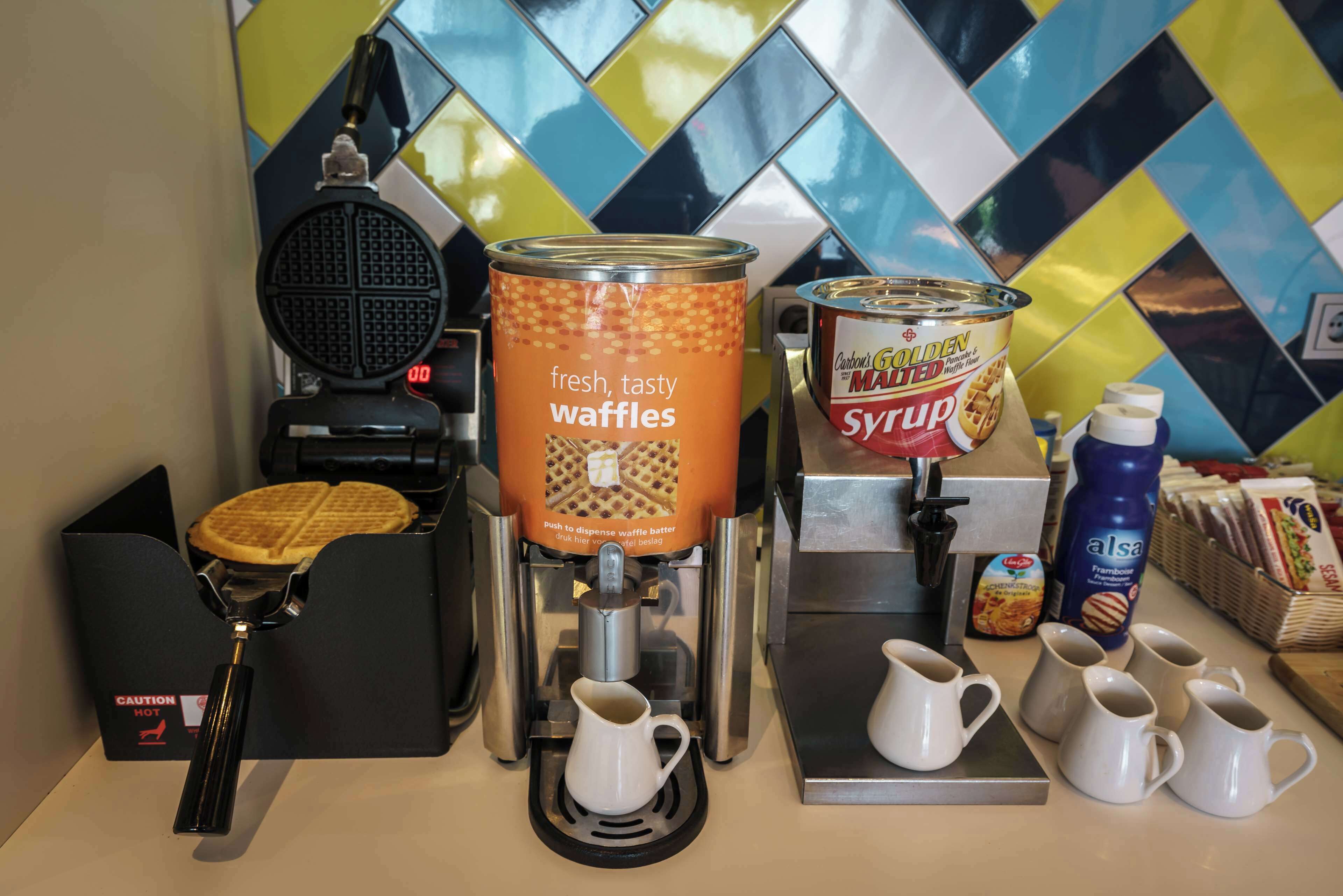 A waffle station with a dispenser, syrup, and waffle maker.