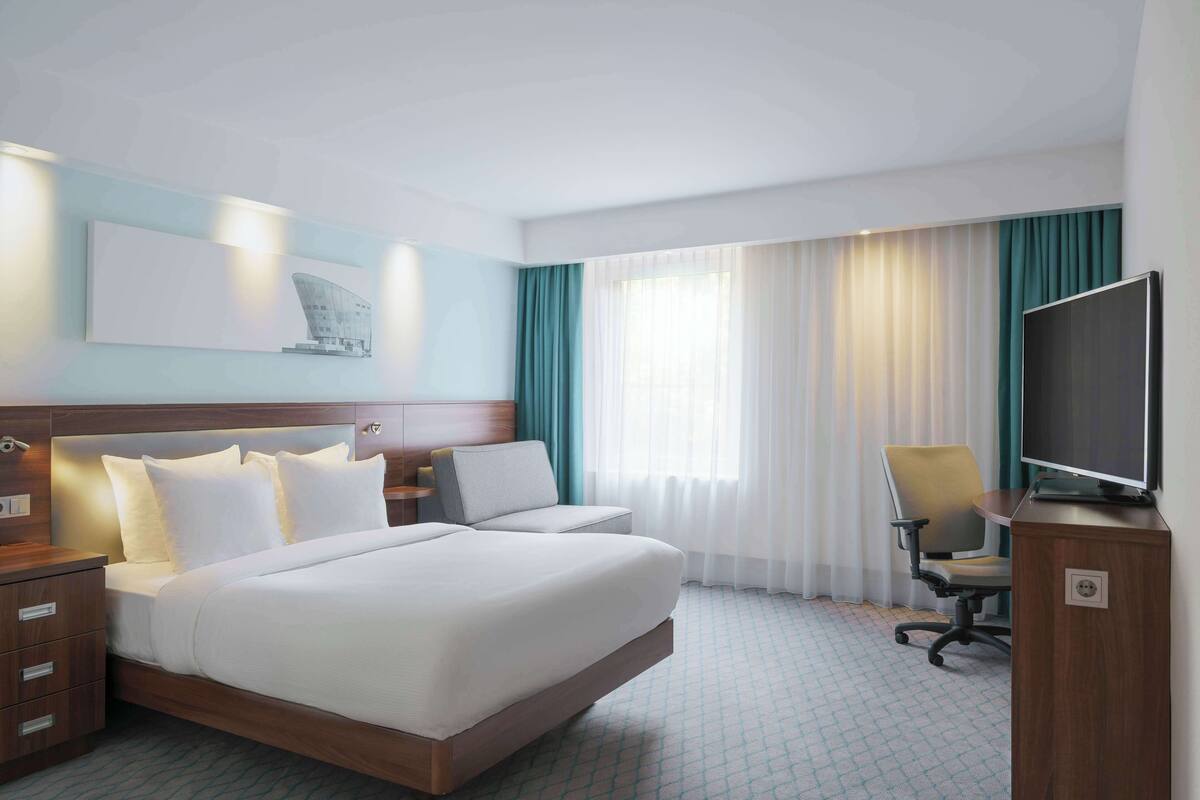 A hotel room with a large bed, a desk with a chair, a TV, and a window with curtains.
