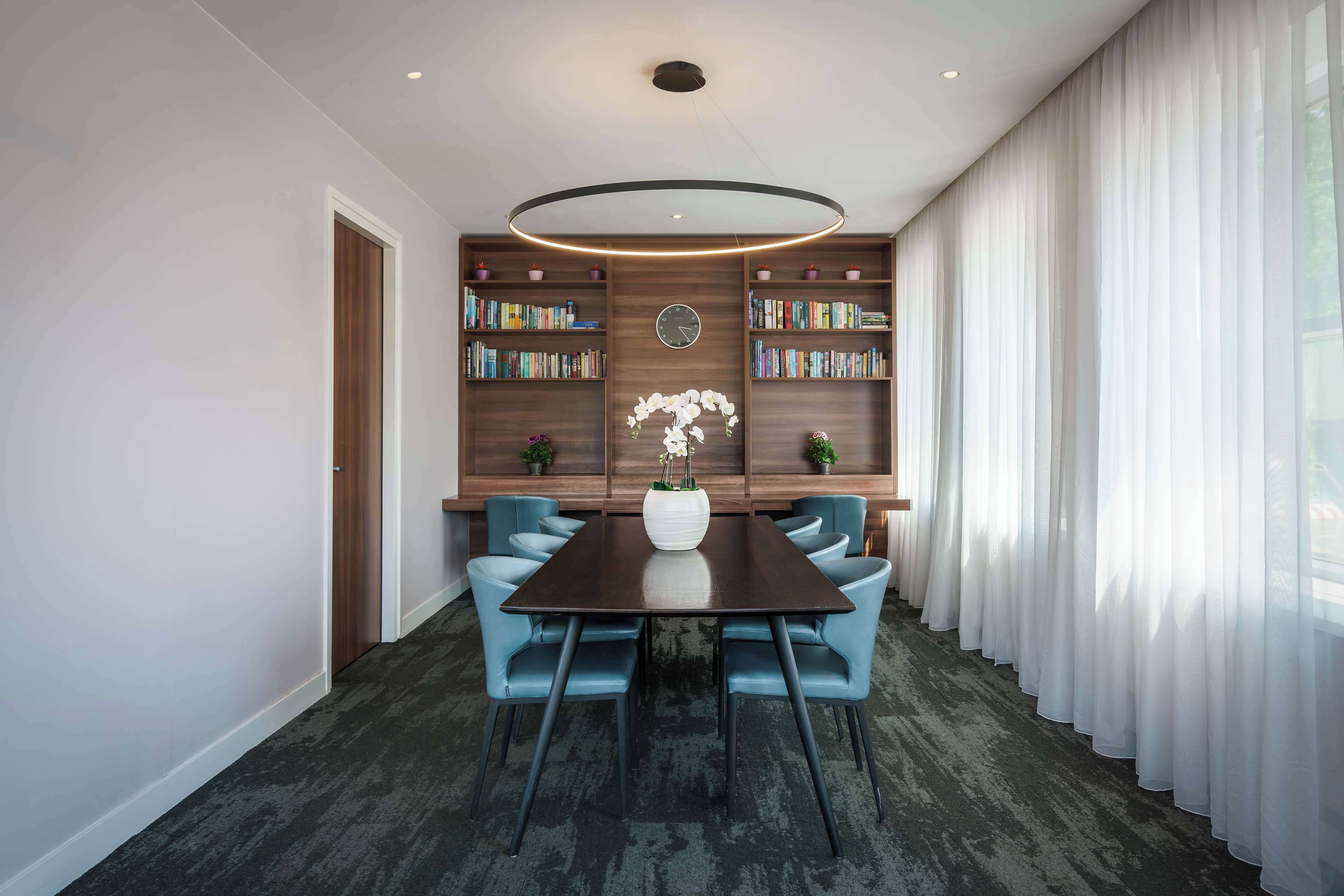 Una sala de conferencias moderna con una mesa de madera oscura, sillas azules y estanterías con libros y objetos decorativos.
