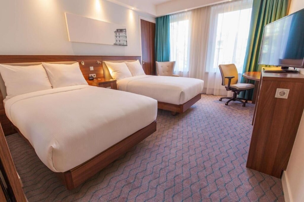 A hotel room with two beds, a desk, a TV, and a window with curtains.