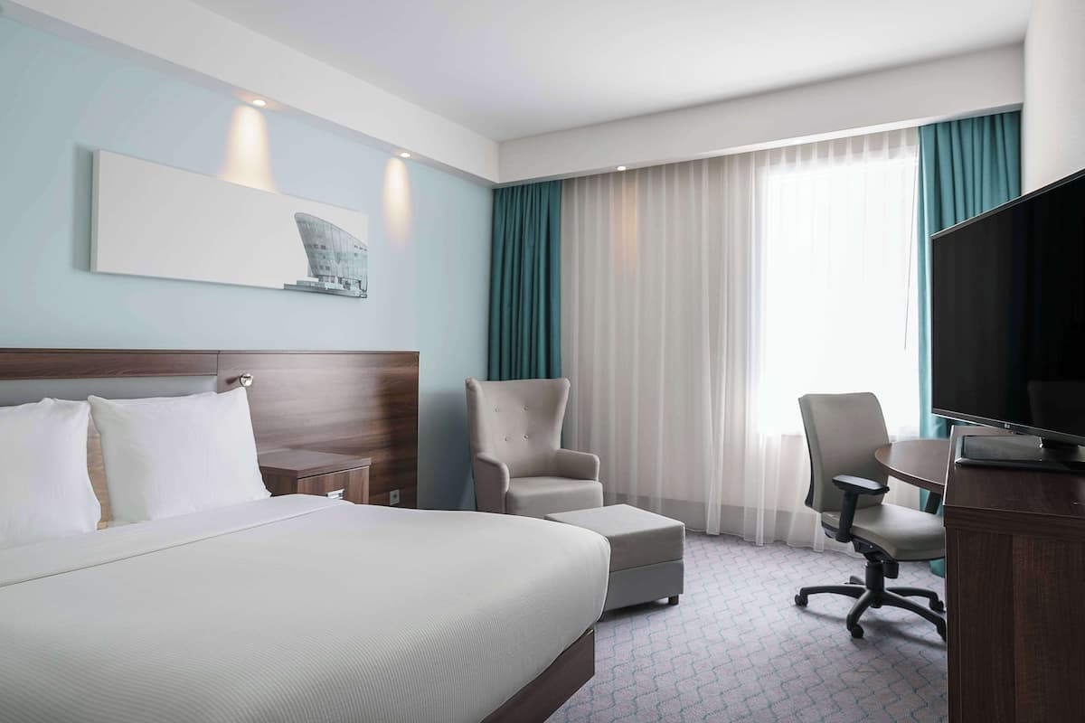 A hotel room with a large bed, two armchairs, a desk with a TV, and a window with curtains.
