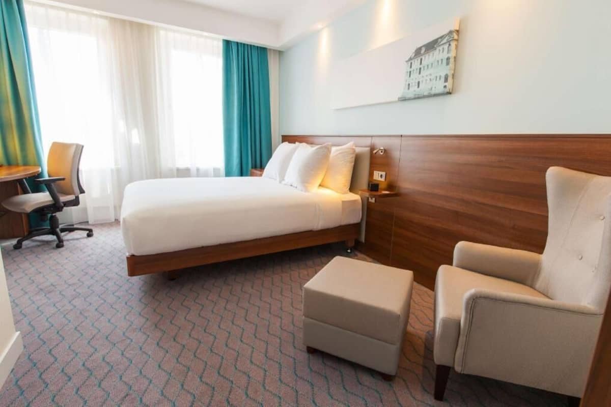 A hotel room with a bed, a desk, a chair, and a small ottoman.