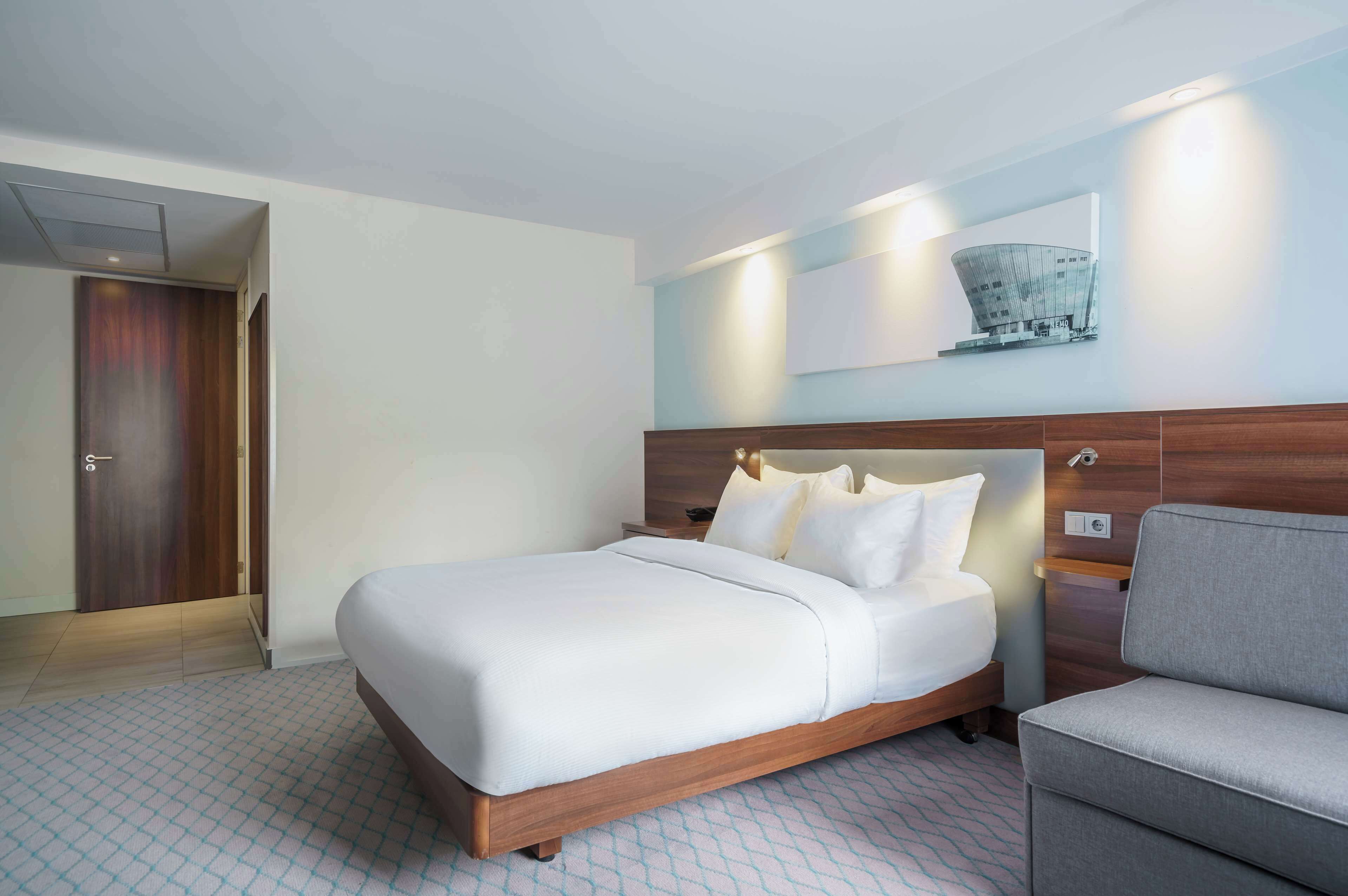 A modern hotel room with a large bed, a grey sofa, a wooden headboard, and a wall-mounted artwork.
