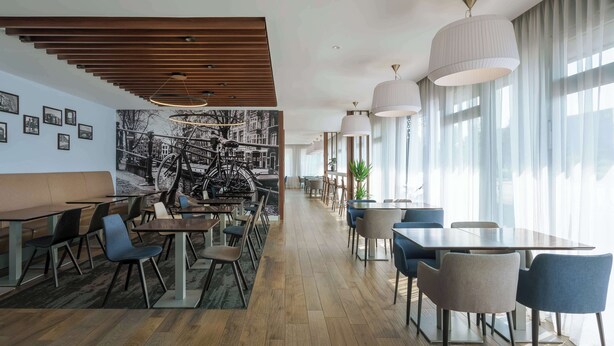 A modern restaurant with wooden flooring, a variety of seating including tables and chairs, and large windows with sheer curtains. - Hampton by Hilton Amsterdam Centre East (Amsterdam)