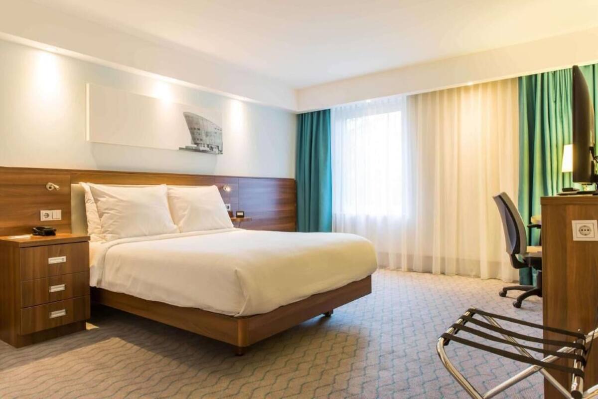 A hotel room with a bed, bedside table, lamp, and a desk with a chair.