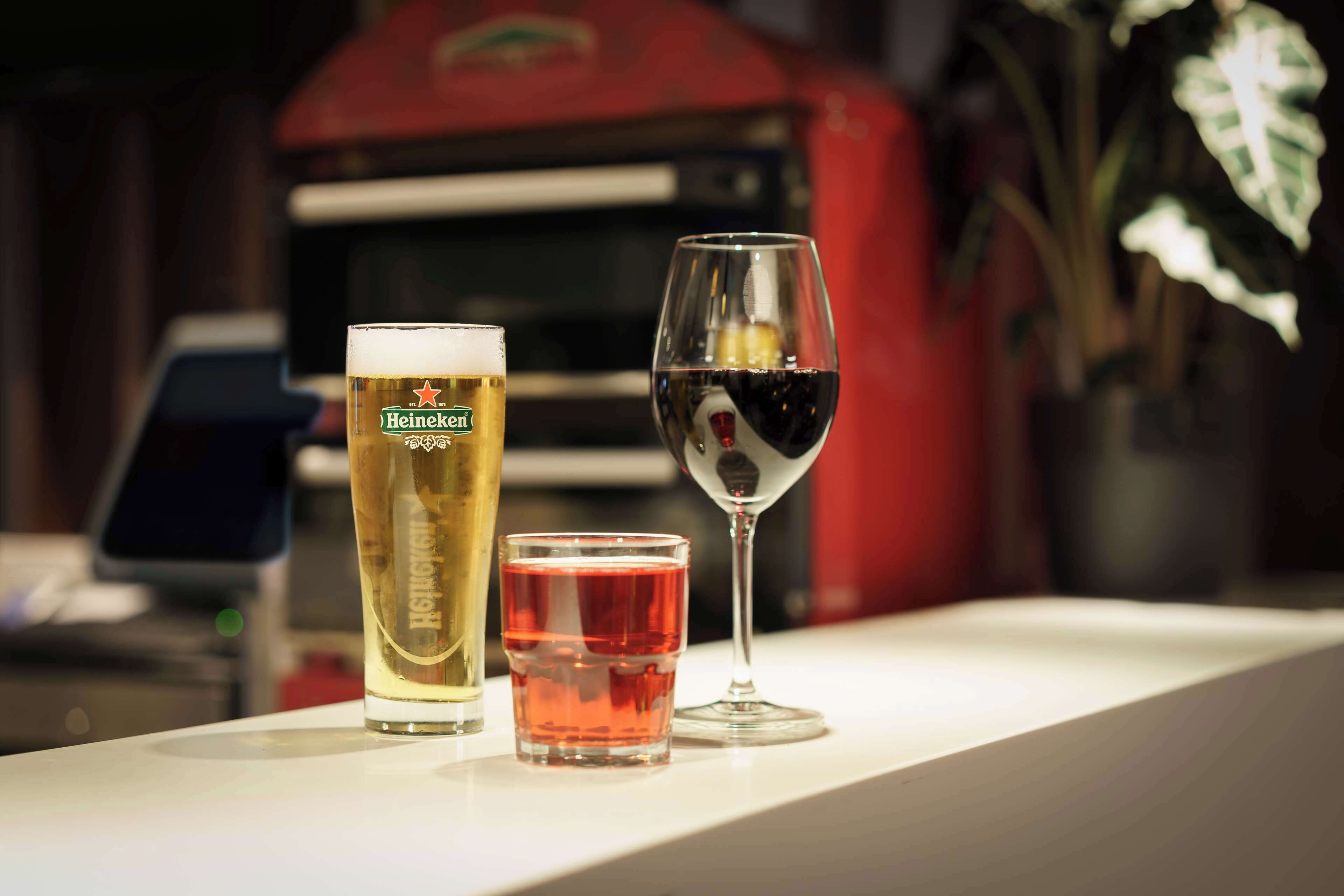 Una cerveza Heineken en vaso, un vaso pequeño con líquido rojo, y una copa de vino con líquido rojo sobre una superficie blanca.