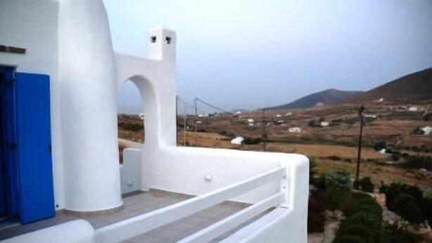 Un edificio blanco con puerta azul, un balcón y vistas a un paisaje rural con casas y colinas.