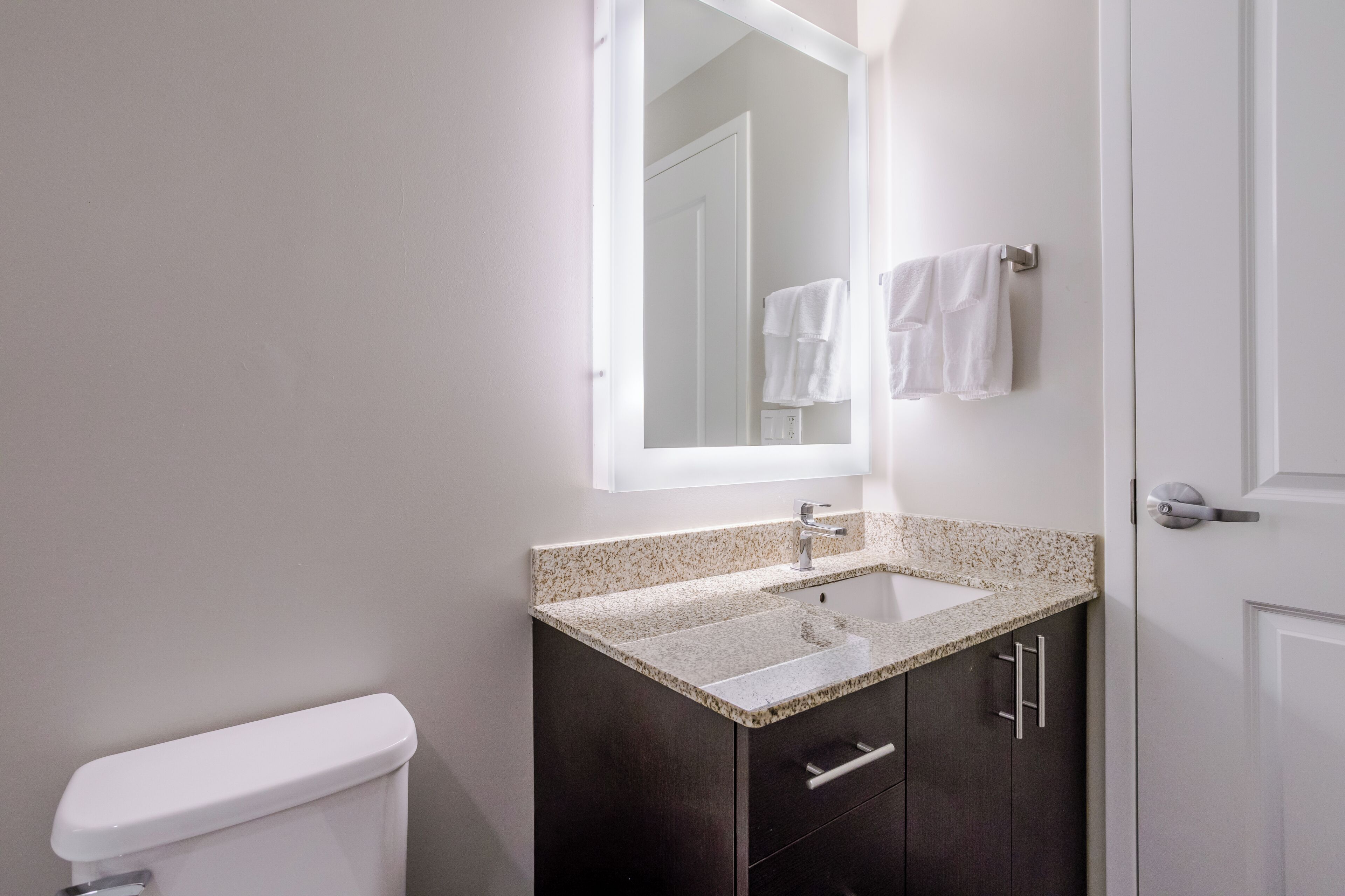 A bathroom with a toilet, a sink with a granite countertop, dark cabinets, and a mirror above the sink.