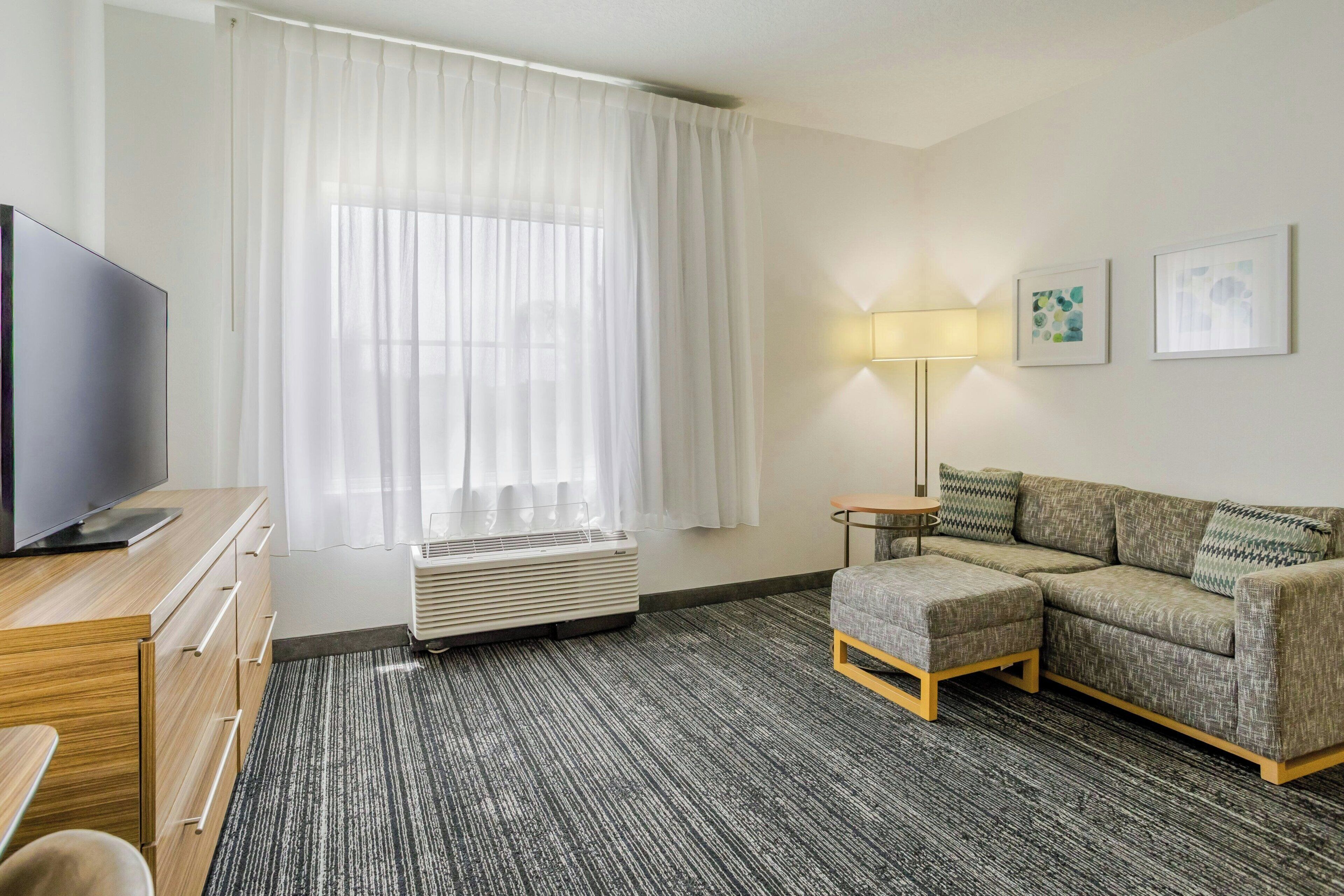 Une chambre d’hôtel équipée d’un téléviseur à écran plat, d’un canapé, d’une petite table, d’une lampe et d’une fenêtre avec des rideaux légers.