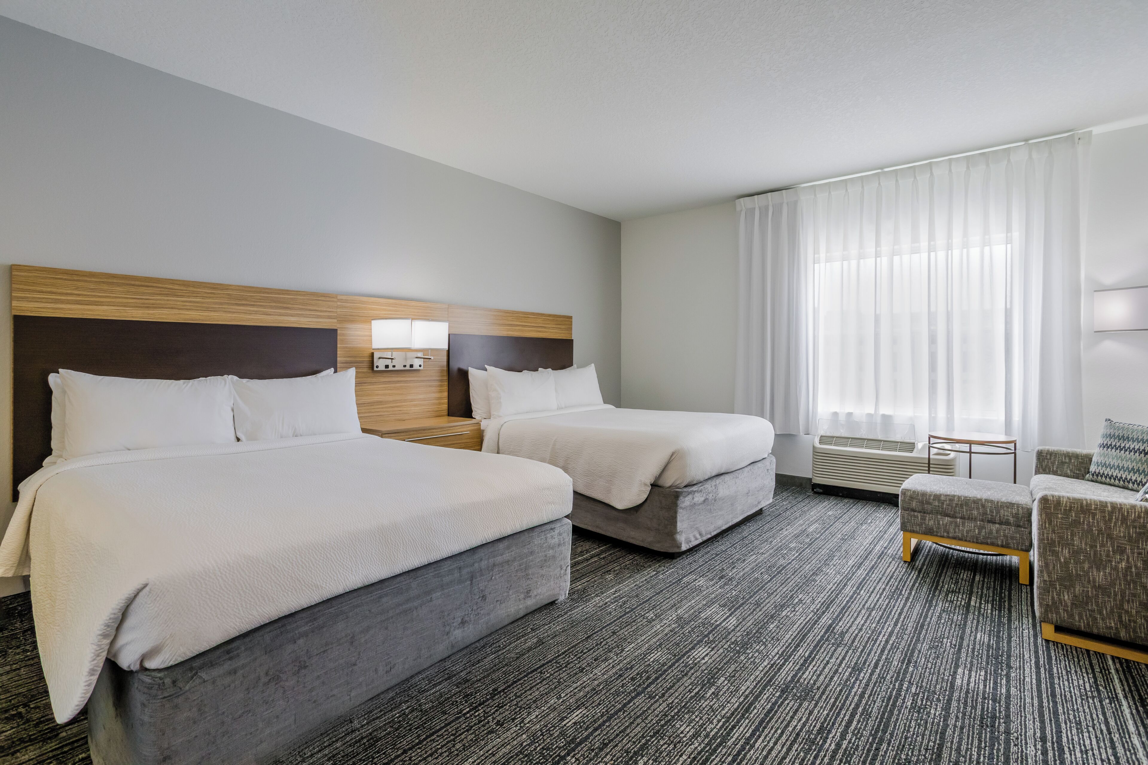 A hotel room with two beds, a small table, a chair, and a large window with curtains.