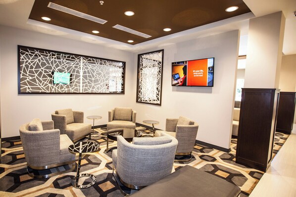 Een moderne hotel lobby met een zithoek met grijze gestoffeerde stoelen en een kleine ronde tafel. Er zijn decoratieve wandpanelen en een flatscreen televisie aan de muur bevestigd.