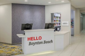 A reception desk with a 'HELLO Boynton Beach' sign, a patterned wall, and a shelf with various items.