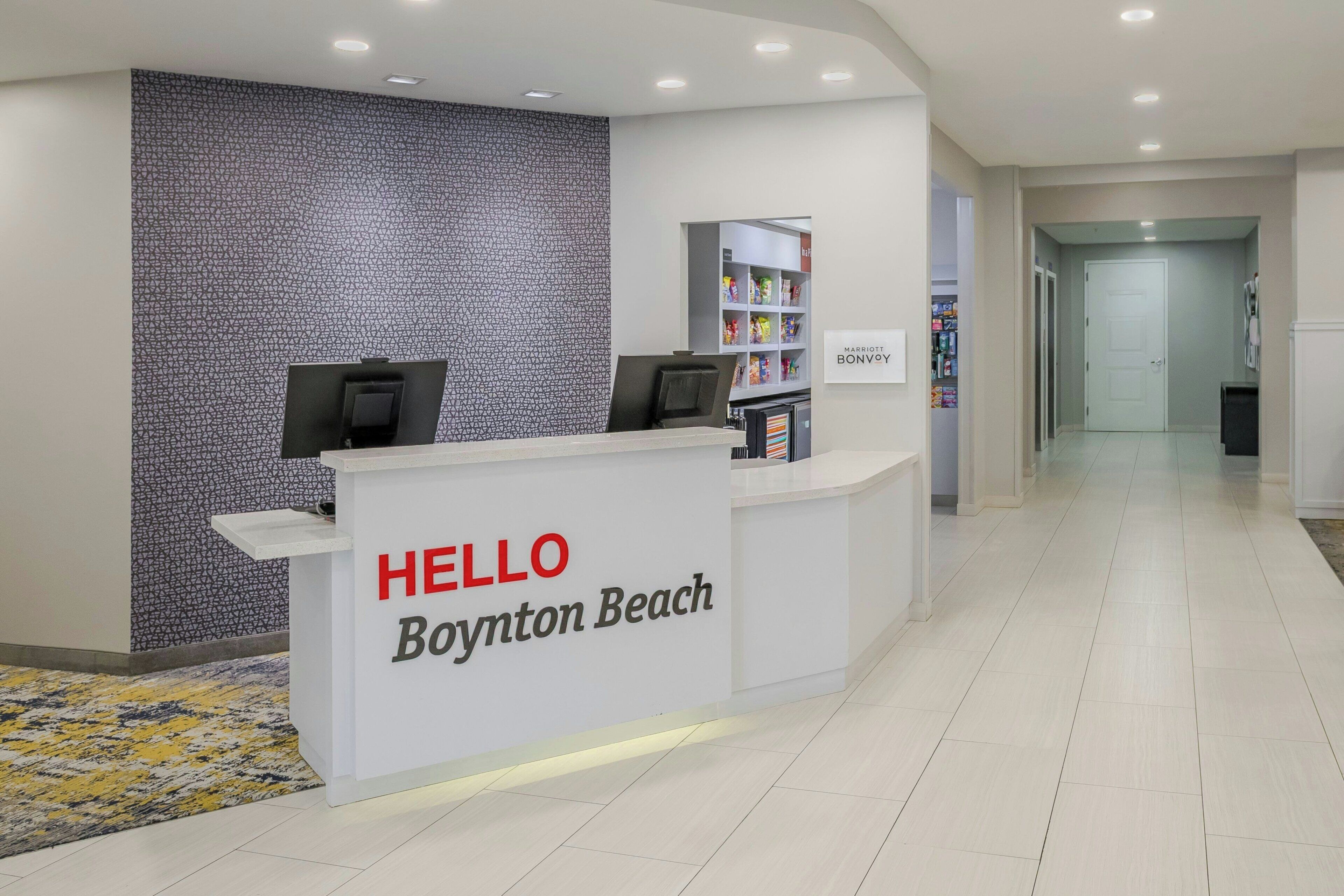 A reception desk with a 'HELLO Boynton Beach' sign, a patterned wall, and a shelf with snacks.