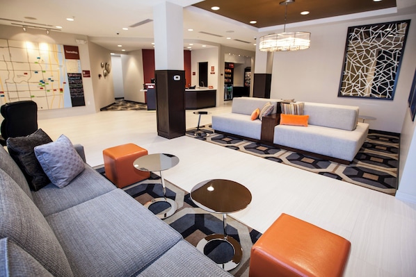Eine moderne Hotelhalle mit einem grauen Sofa, einem orangefarbenen Hocker und einem Couchtisch. Im Hintergrund ist eine Rezeption erkennbar und an der Wand hängt eine Karte.