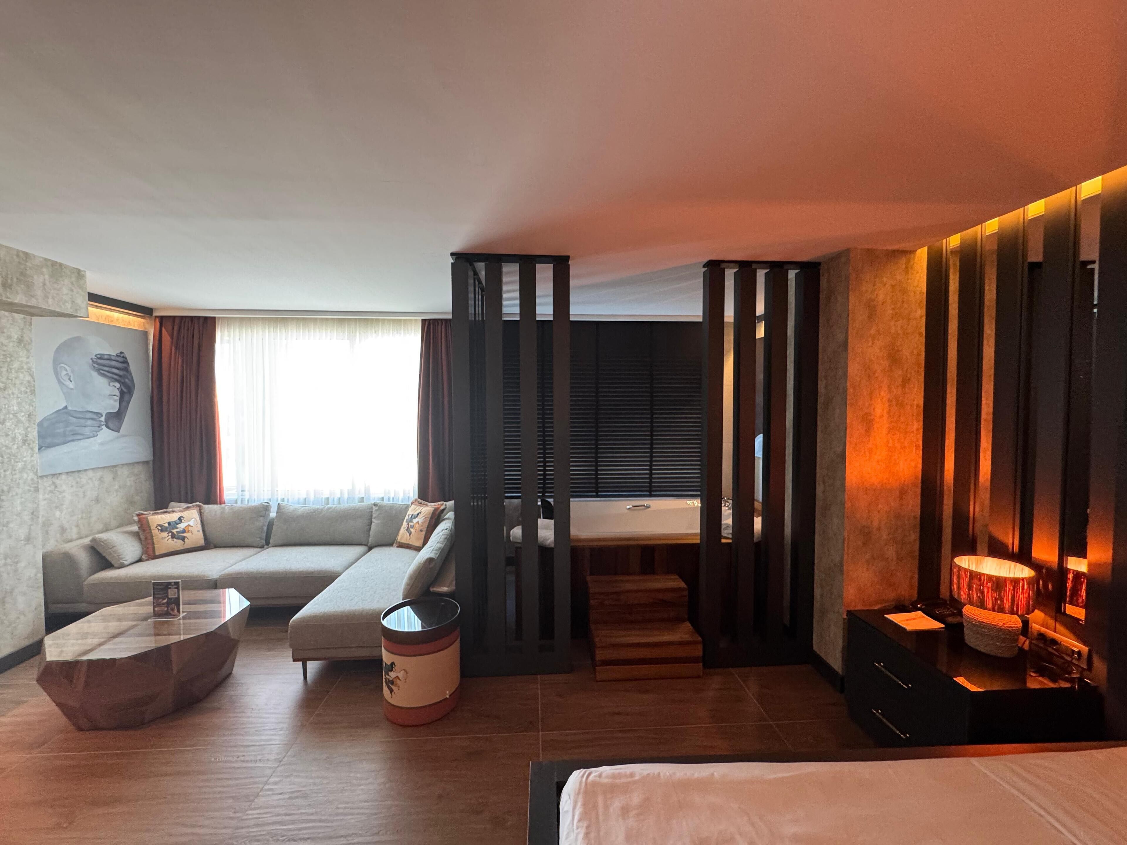 Una camera d'albergo moderna con divano, tavolino da caffè, un piccolo tavolino di servizio, un letto, un bagno con vasca e un'ampia finestra con tende.