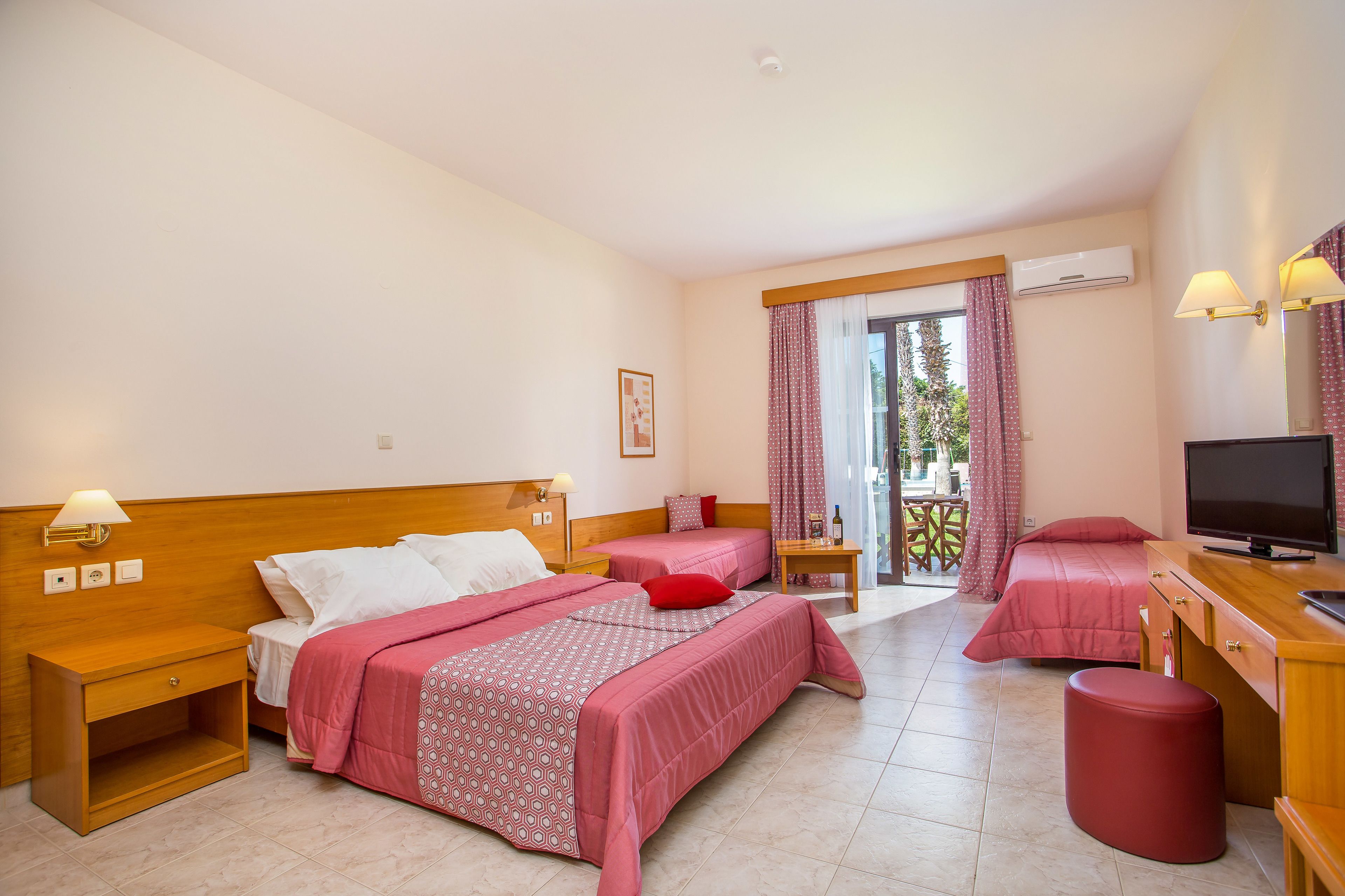 A hotel room with two beds, a TV, a wooden nightstand, a red stool, and a balcony with curtains.
