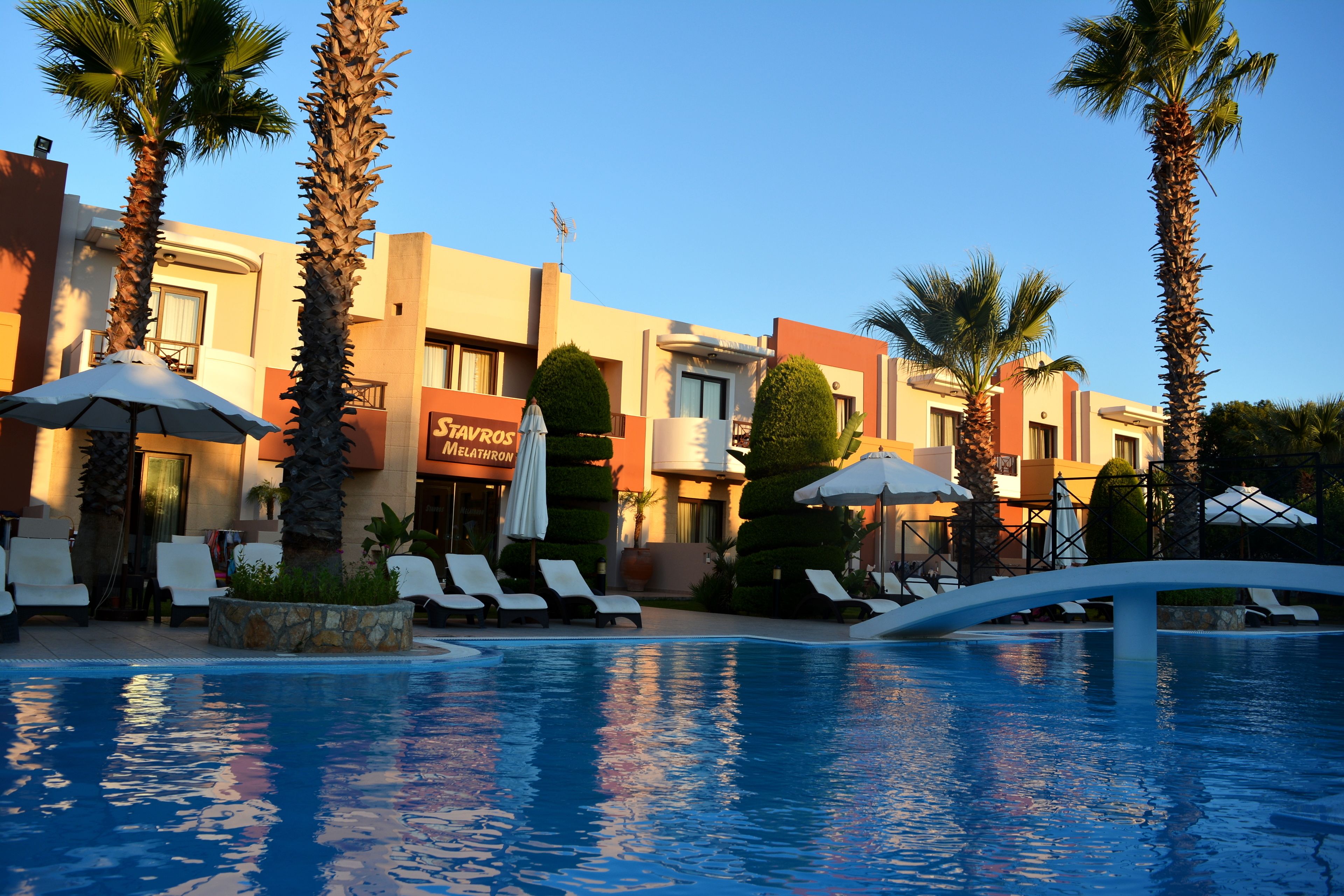 Zona de piscina de hotel con tumbonas, palmeras y un cielo azul despejado.