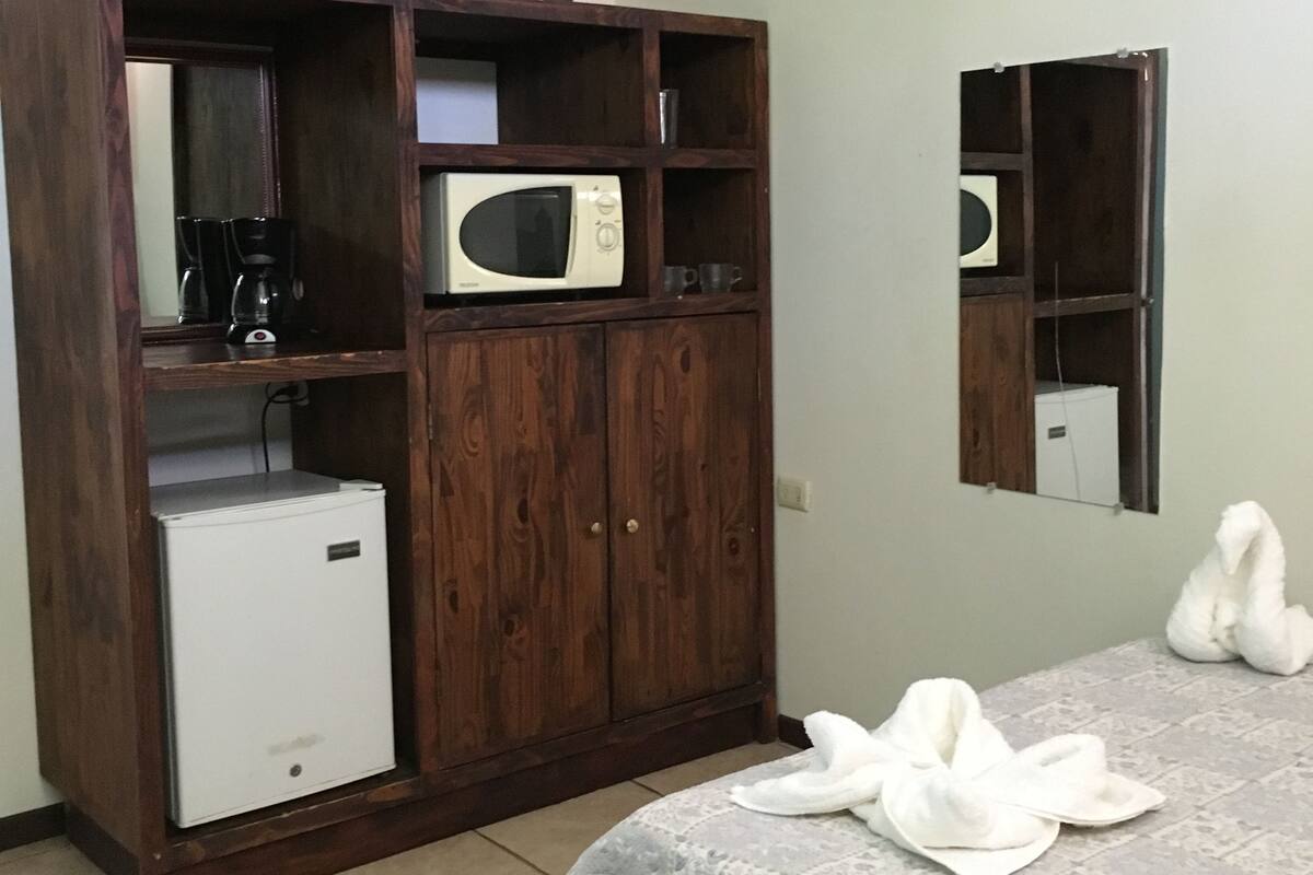 A room with a wooden cabinet containing a microwave, a small fridge, and a television. There is a table with a tablecloth and towels.
