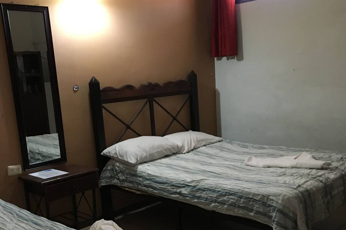 A room with two beds, a mirror, a small table, and a window with a red curtain.