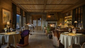 A cozy, rustic dining area with wooden beams, stone walls, and leather furniture. Tables are set with wine glasses and plates, and a fireplace adds warmth.