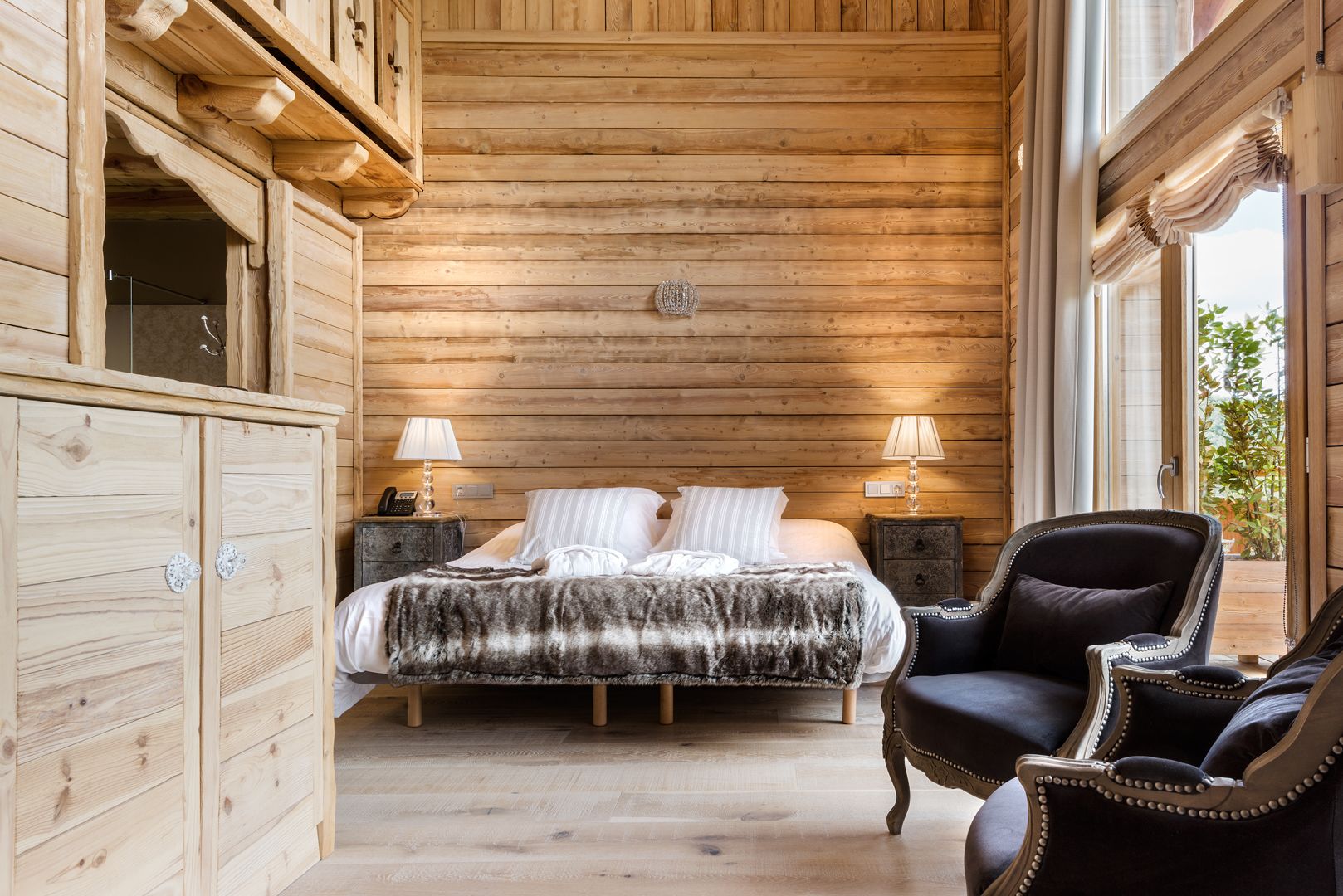 A wooden cabin bedroom with a bed, two bedside lamps, a chair, and a cabinet.