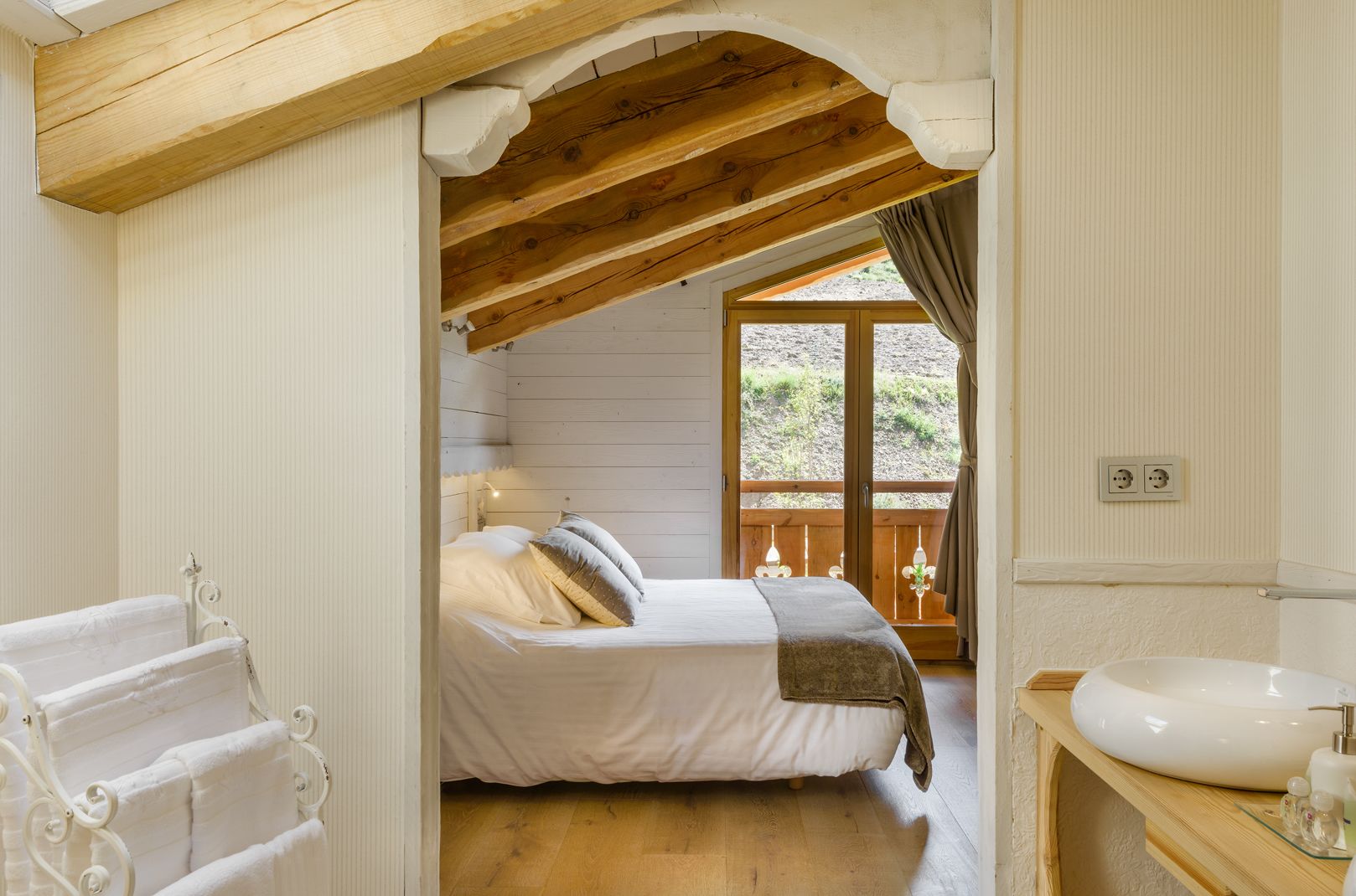 Un dormitorio con techo de madera, una cama con ropa blanca, una ventana con vistas al exterior y un baño con lavabo y espejo.