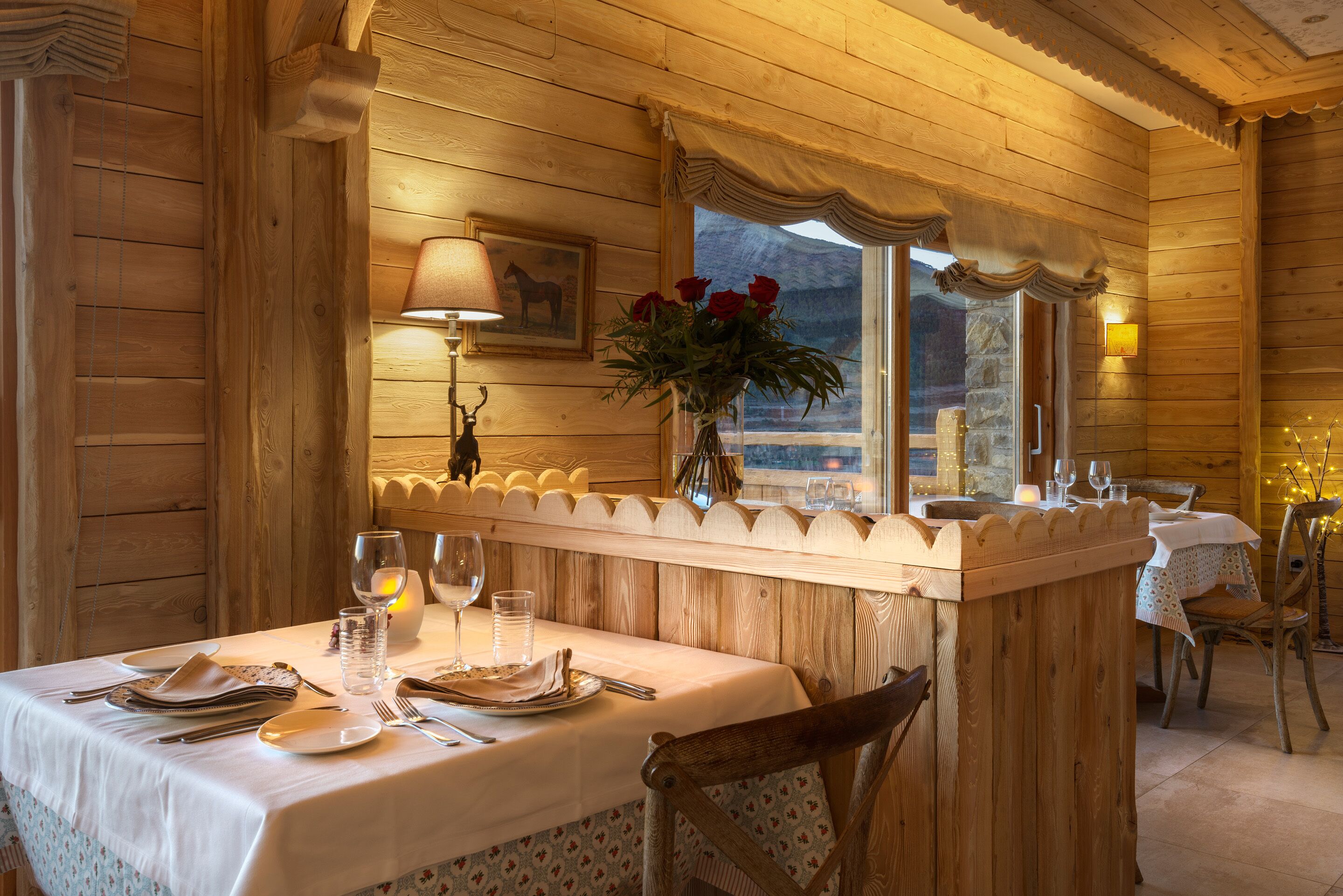 Interior de uma cabana de madeira com uma mesa de jantar posta para dois, um vaso com flores e uma janela com cortinas.