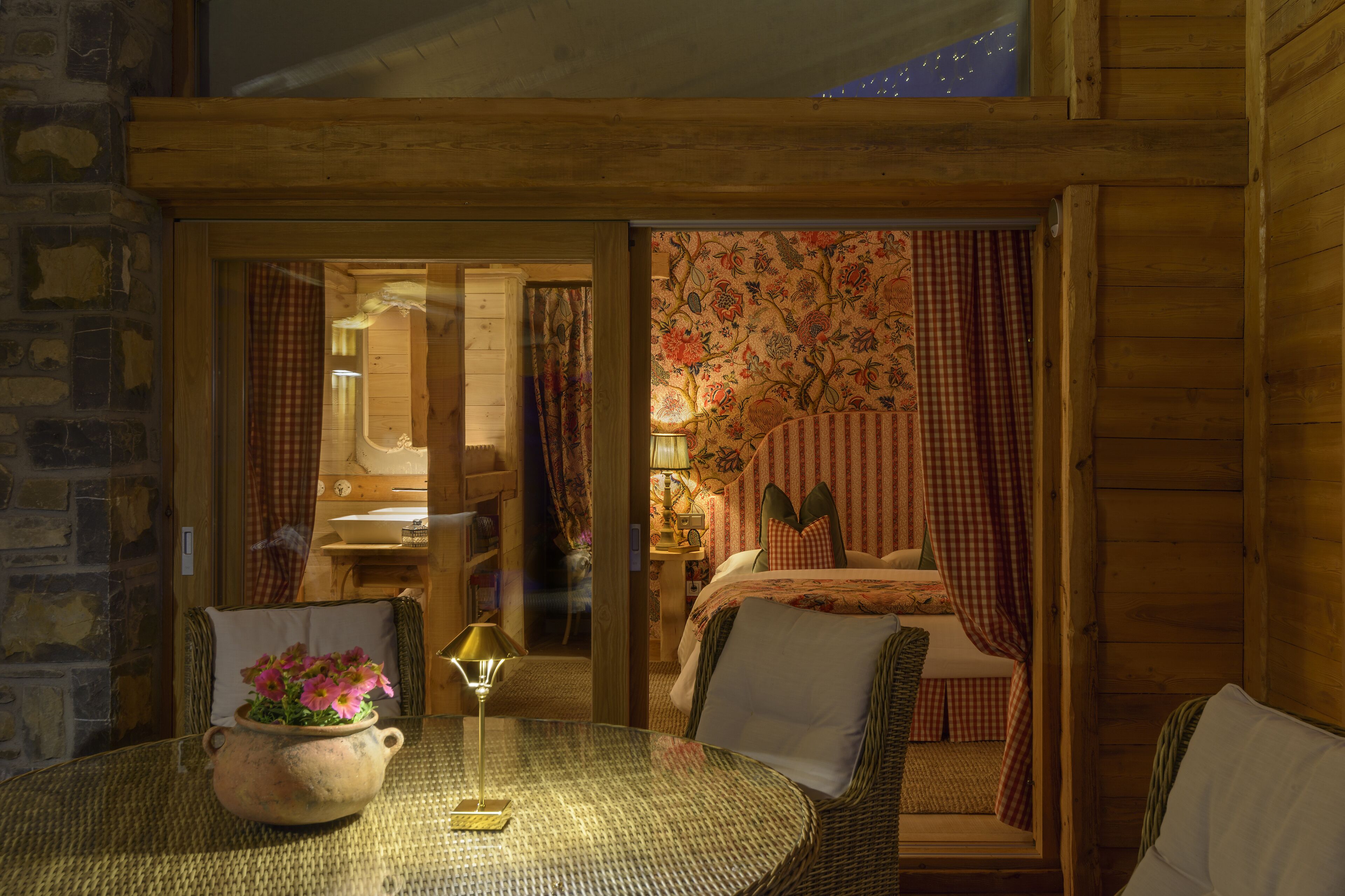 Een knus ingerichte kamer met een rieten stoel, een tafel met een bloempot, een bed met geruit gordijn en een houten plafond.