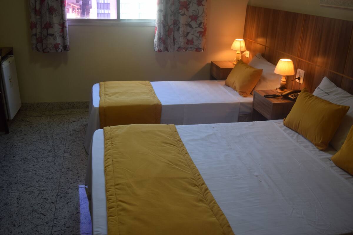 Et hotelværelse med to senge, hver med et gult sengetæppe, et natbord med en lampe, og et vindue med blomstrede gardiner.