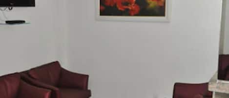 Una habitación con techo blanco, un televisor de pantalla plana montado en la pared y un cuadro con flores rojas. Hay tres sillas rojas dispuestas en la habitación.