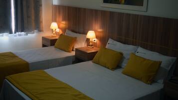Quarto de hotel com duas camas, cada uma com colcha branca e almofadas amarelas. Há duas mesinhas de cabeceira com abajures, e um quadro é visível na parede acima das camas.