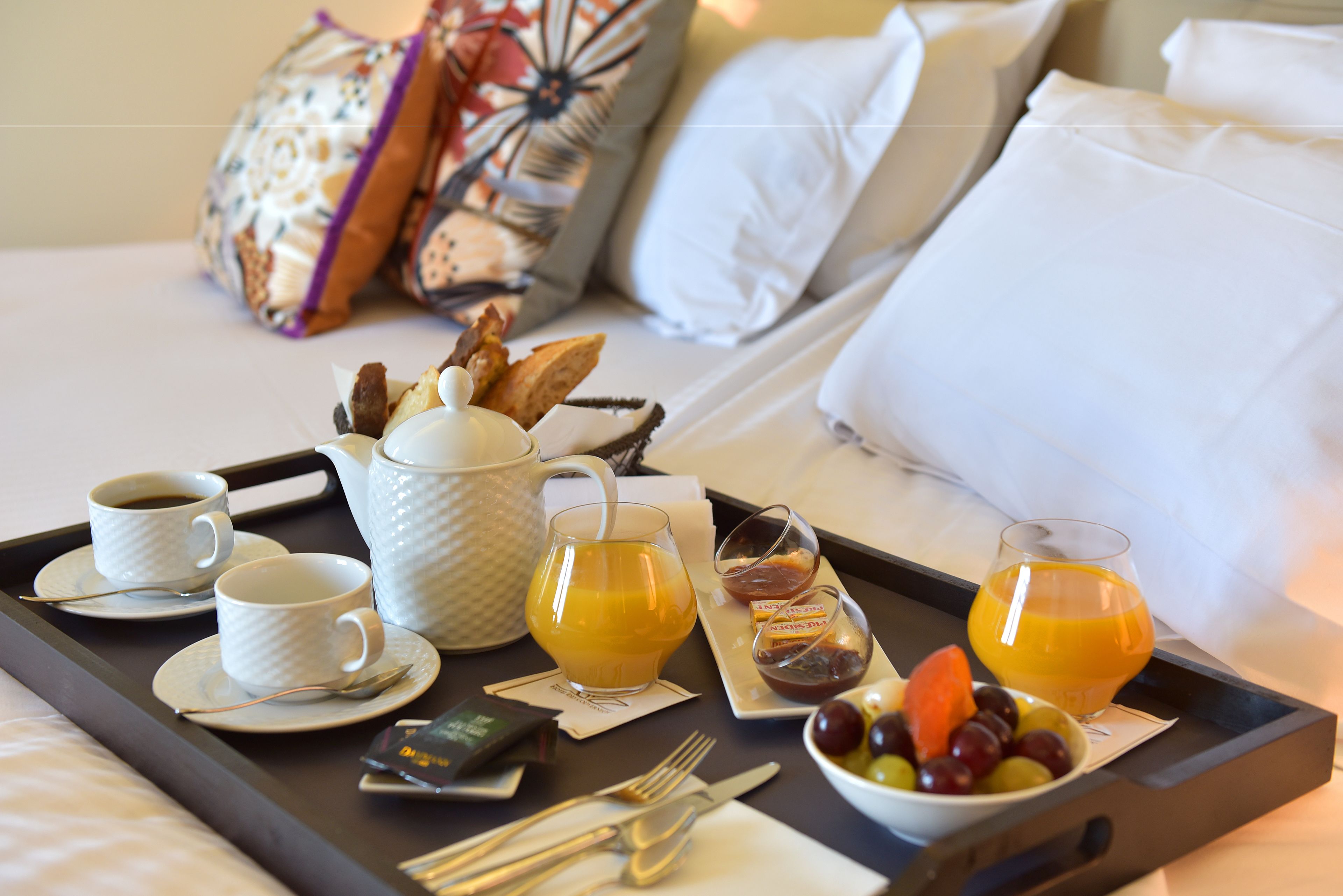 A breakfast tray with coffee cups, orange juice, and a bowl of fruit on a bed with pillows.