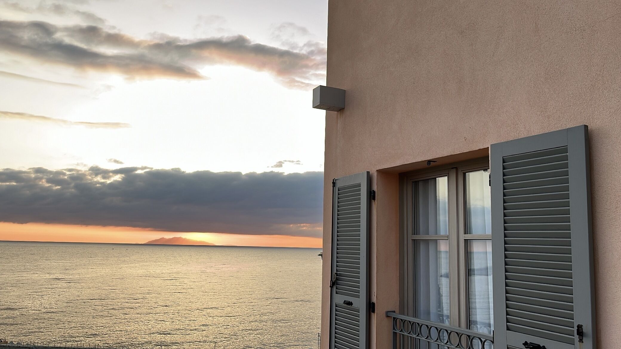 A balcony with a view of the sunset over the sea, a window with shutters, and a wall-mounted device.