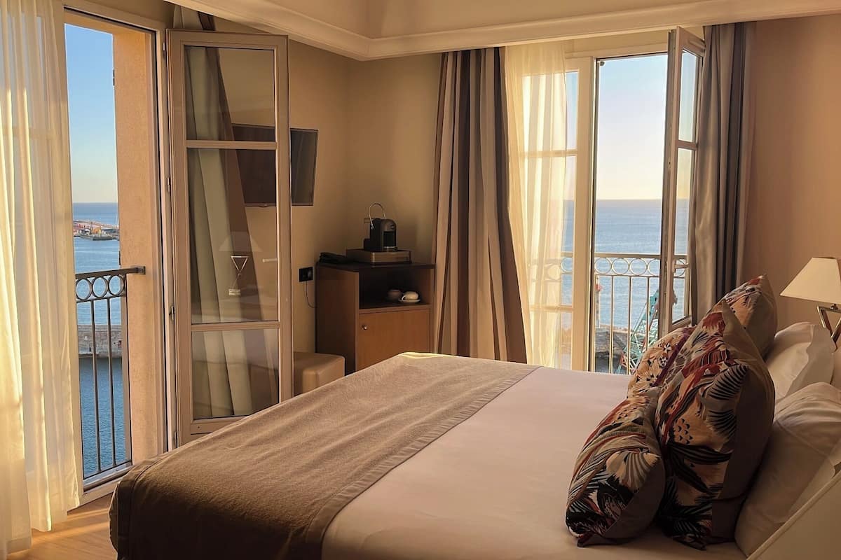 A hotel room with a bed, a nightstand, a small cabinet, and a view of the sea through large windows.