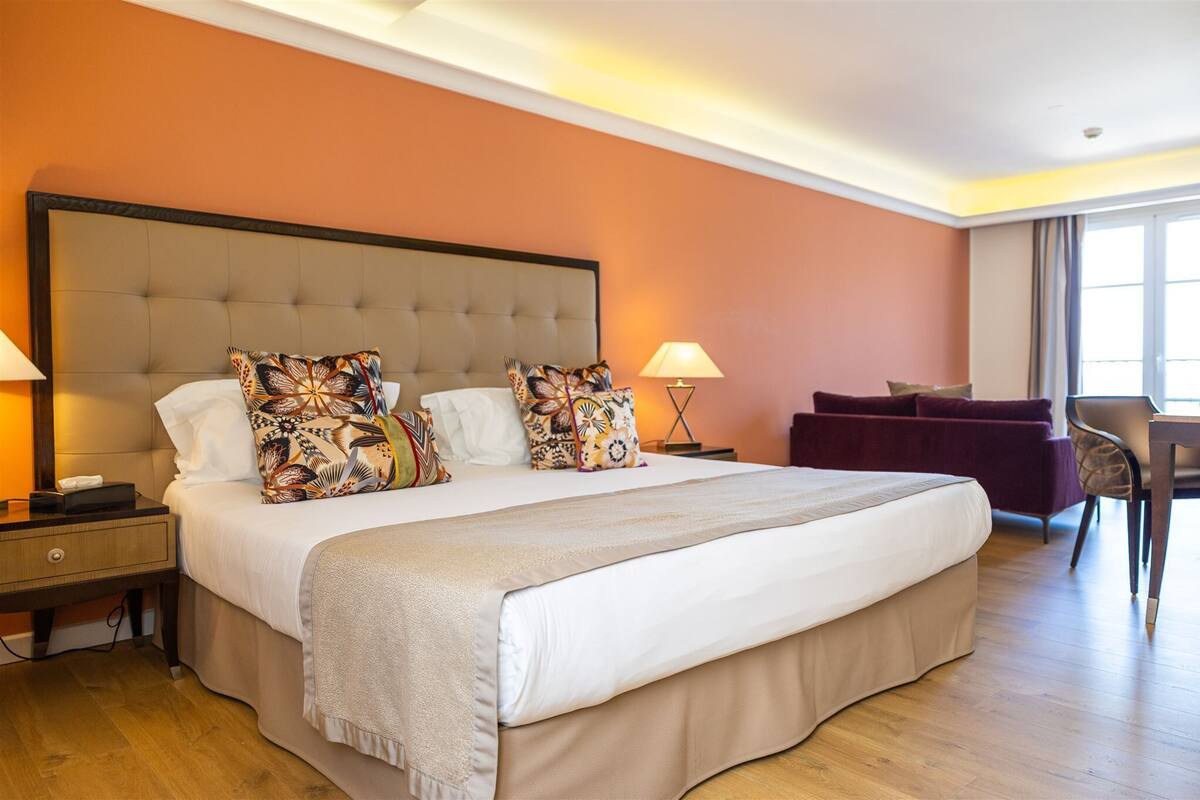 A hotel room with a large bed, two bedside tables, a nightstand, a purple armchair, and a desk with a chair.