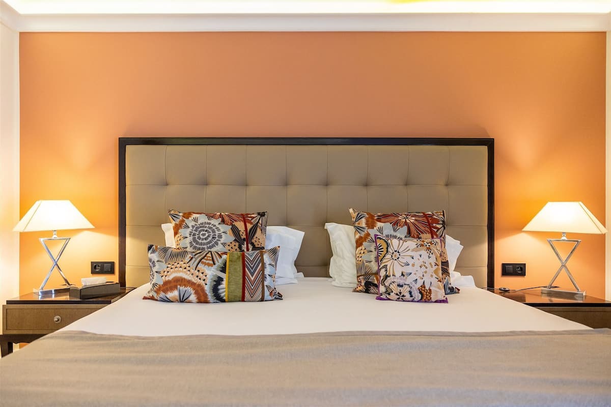 A neatly made bed with multiple patterned pillows, two bedside lamps, and a nightstand with a phone and a small box.