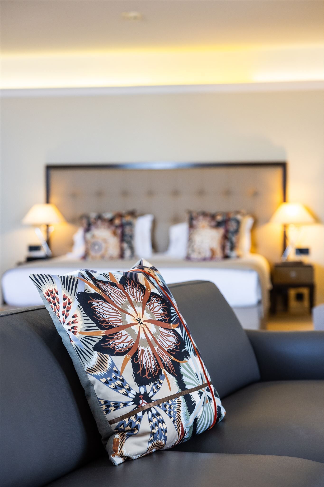 A modern hotel room with a large bed, two bedside lamps, and a decorative pillow on a dark sofa.