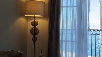 Ein Hotelzimmer mit Bett, Tisch mit Lampe, einer Flasche und Gläsern, und Meerblick vom Balkon.