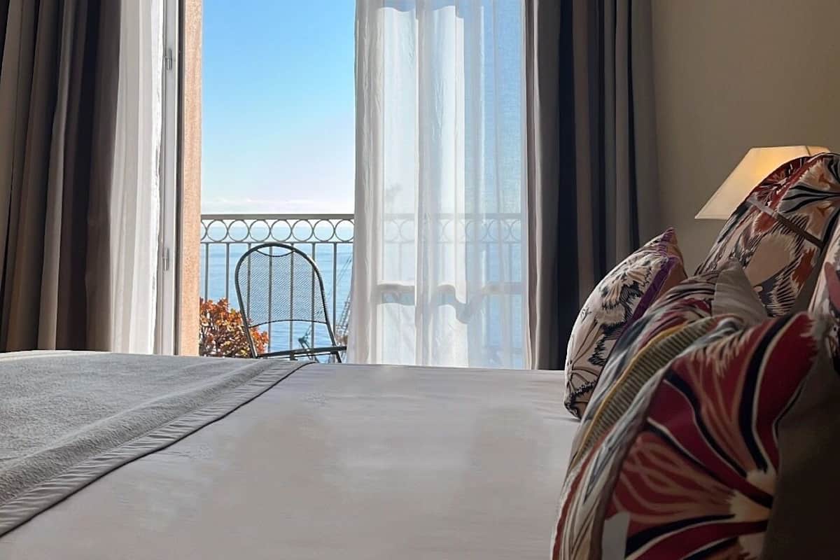 A hotel room with a large bed, a balcony with a view of the sea, and a chair outside.