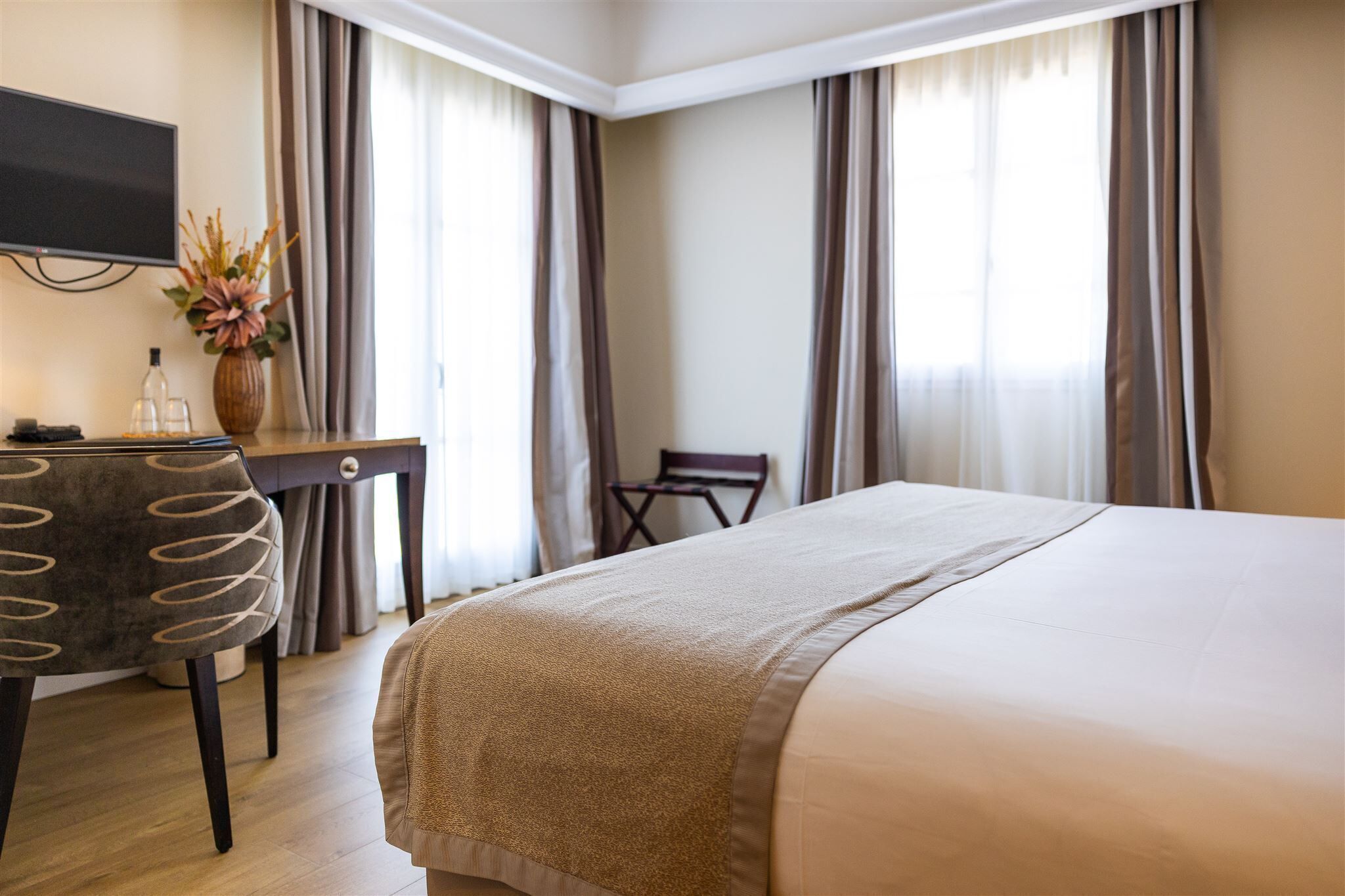 A hotel room with a large bed, a desk with a chair, a television, and a vase with flowers.