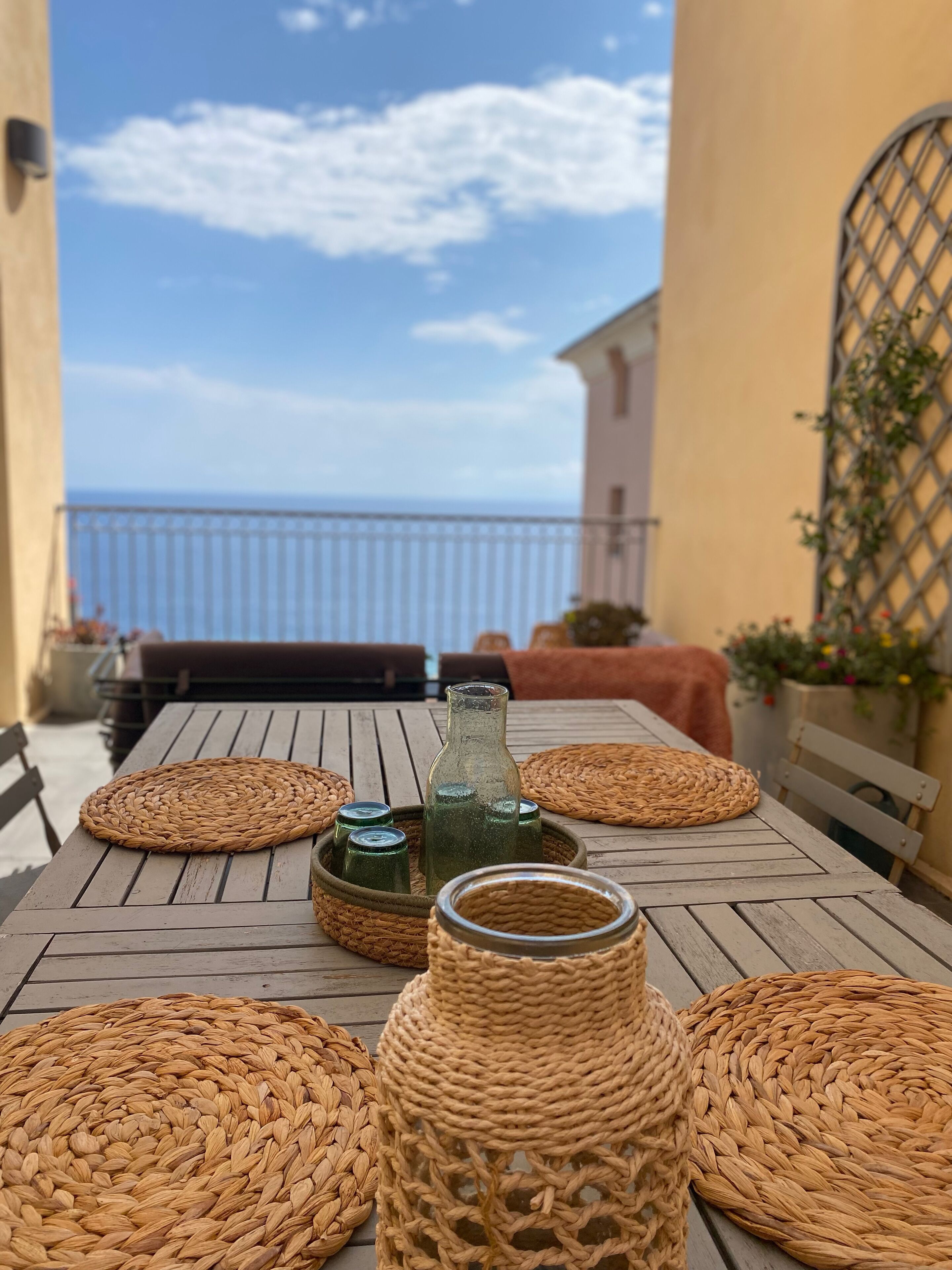 Uma mesa de madeira com jogos americanos trançados, uma garrafa e frascos, numa varanda com vista para o mar.