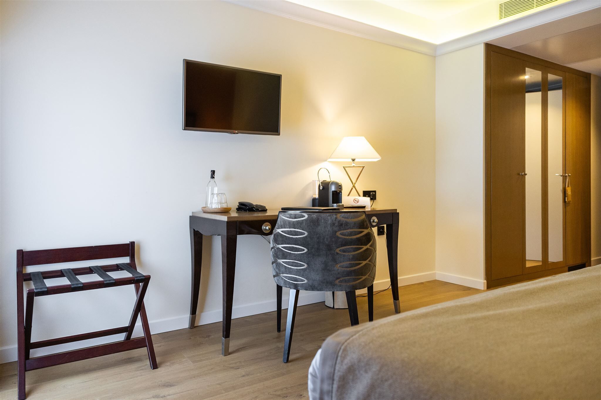 Quarto de hotel com uma cama, uma escrivaninha com cadeira, televisão, luminária e guarda-roupa.
