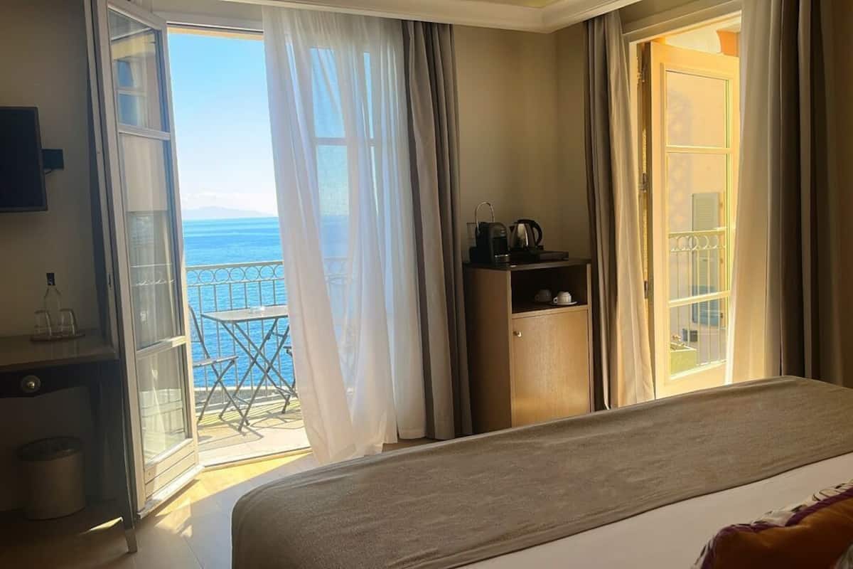 A hotel room with a bed, a small desk, a minibar, and a balcony with a view of the sea.