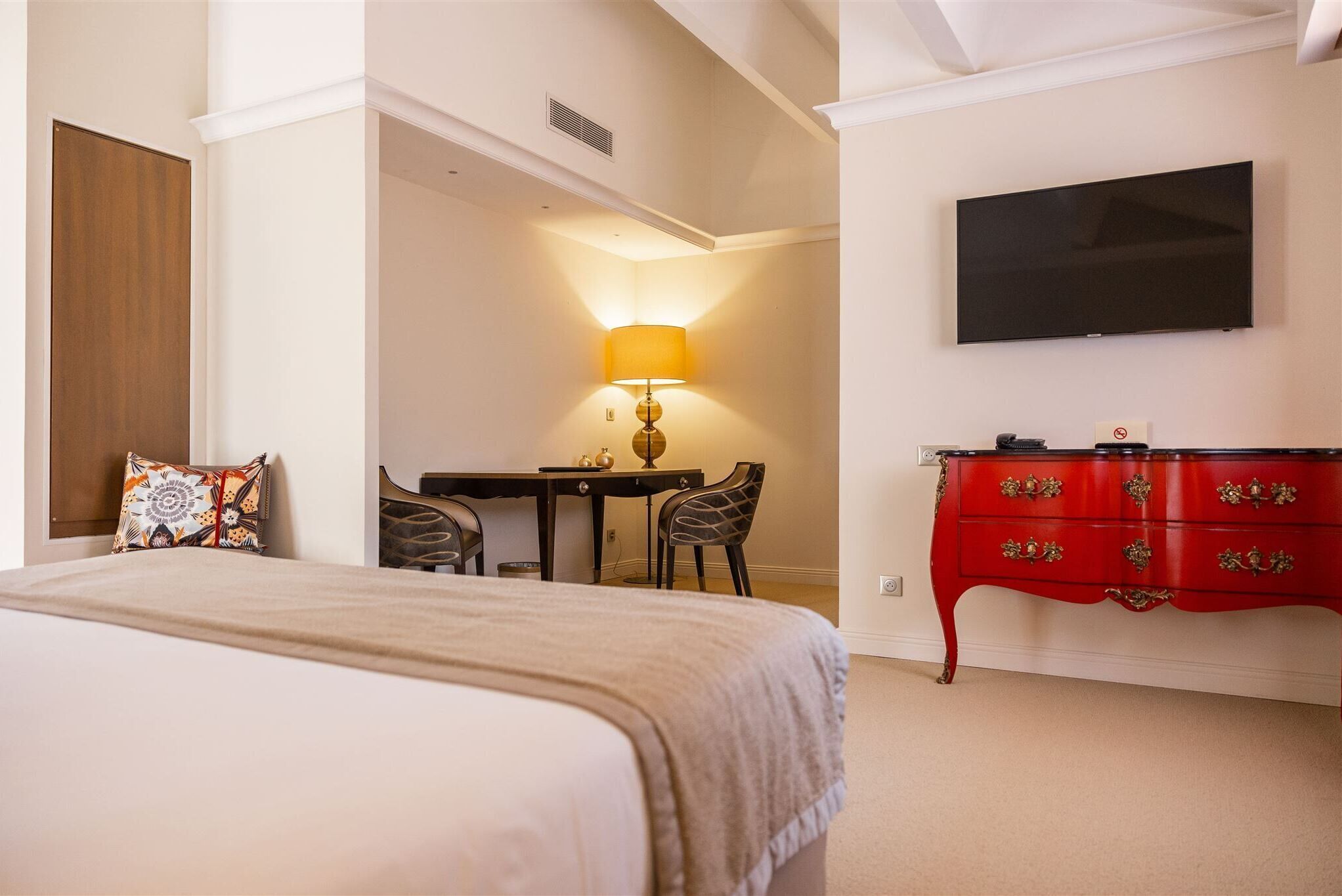 A hotel room with a bed, a desk with a lamp, a TV, and a red dresser.