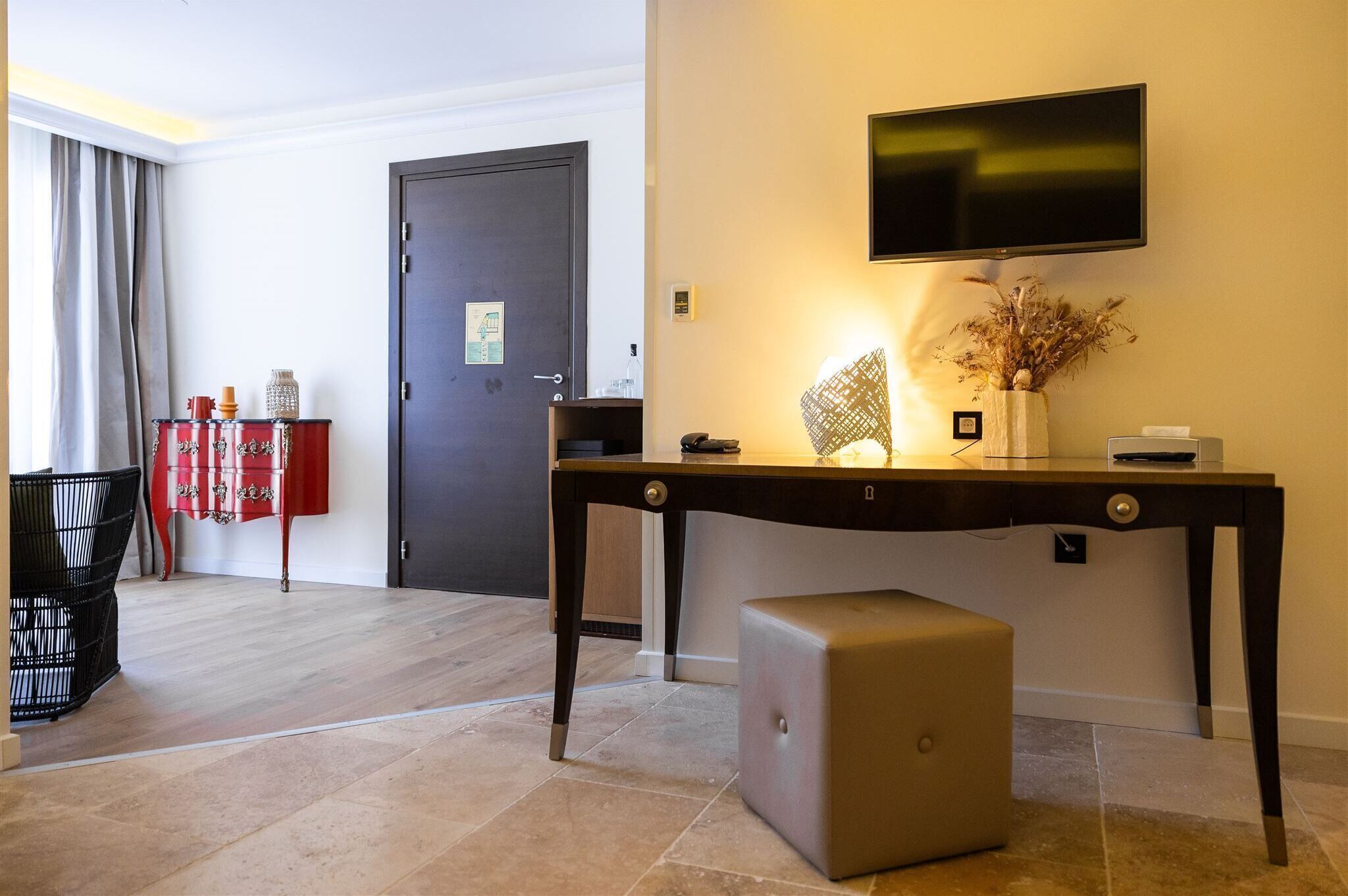 Habitación de hotel con un escritorio de madera, un televisor montado en la pared, una mesita con un jarrón y un armario rojo.