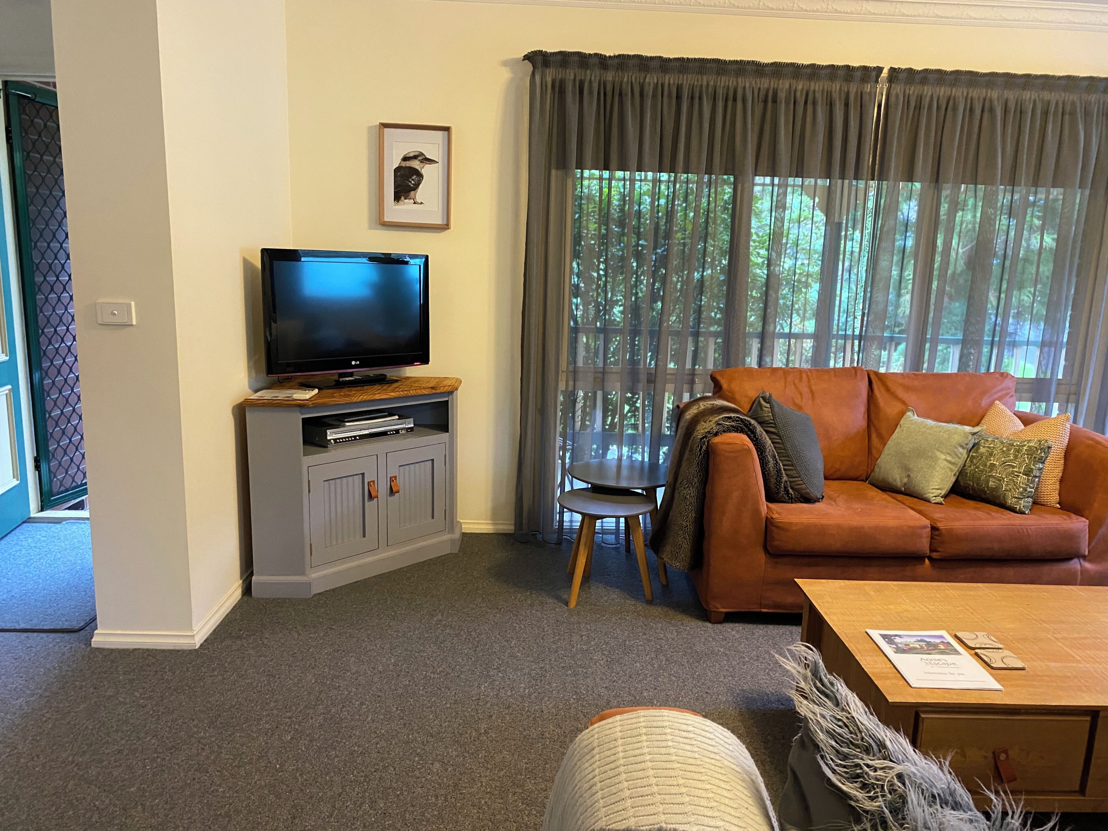 A living room with a television on a cabinet, a brown sofa with cushions, a small table, and a window with curtains.