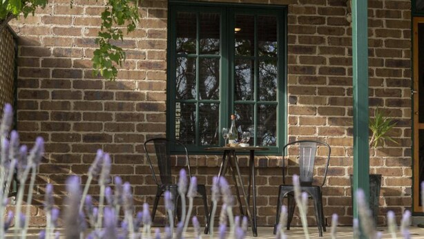 A brick building with a green-framed window, a table with chairs, and lavender plants in the foreground. - Battunga Cottages (Watervale)