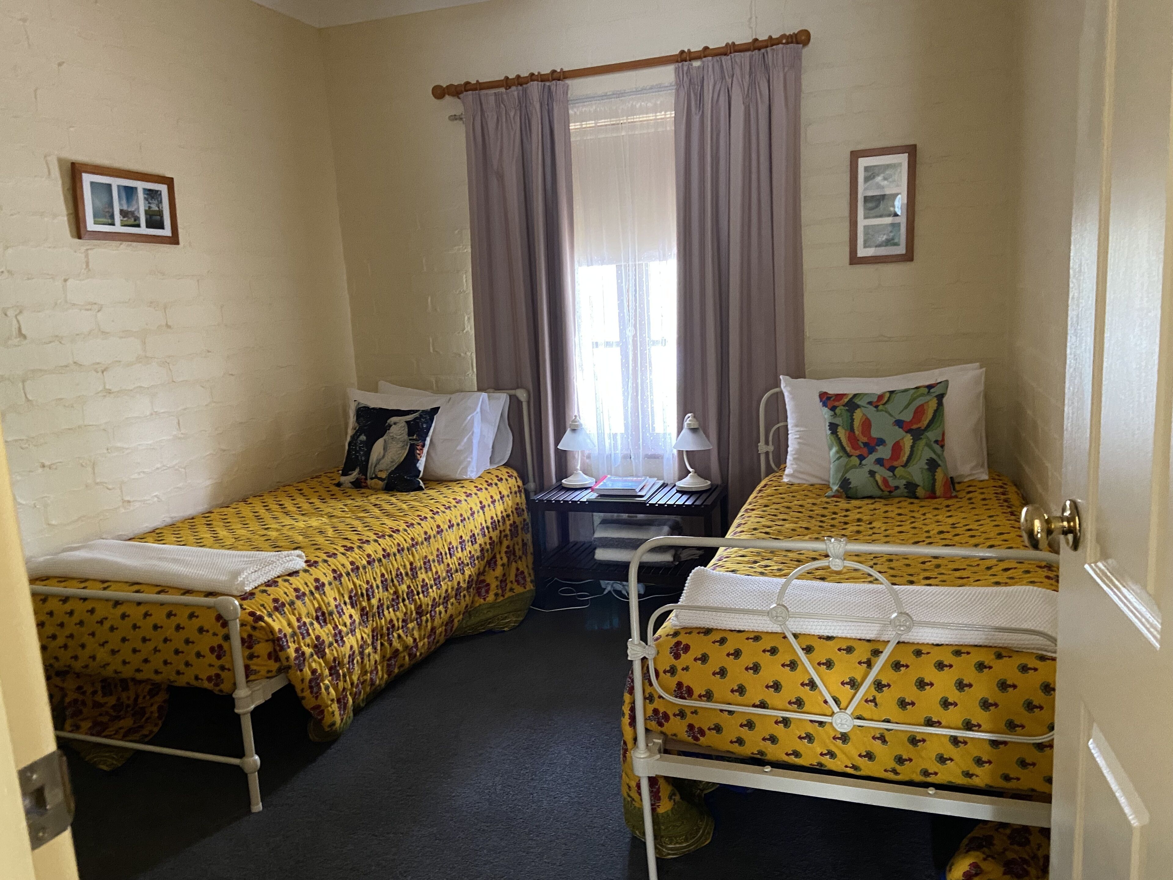 A small, compact room with two single beds, a desk with a chair, a window with curtains, and a door.