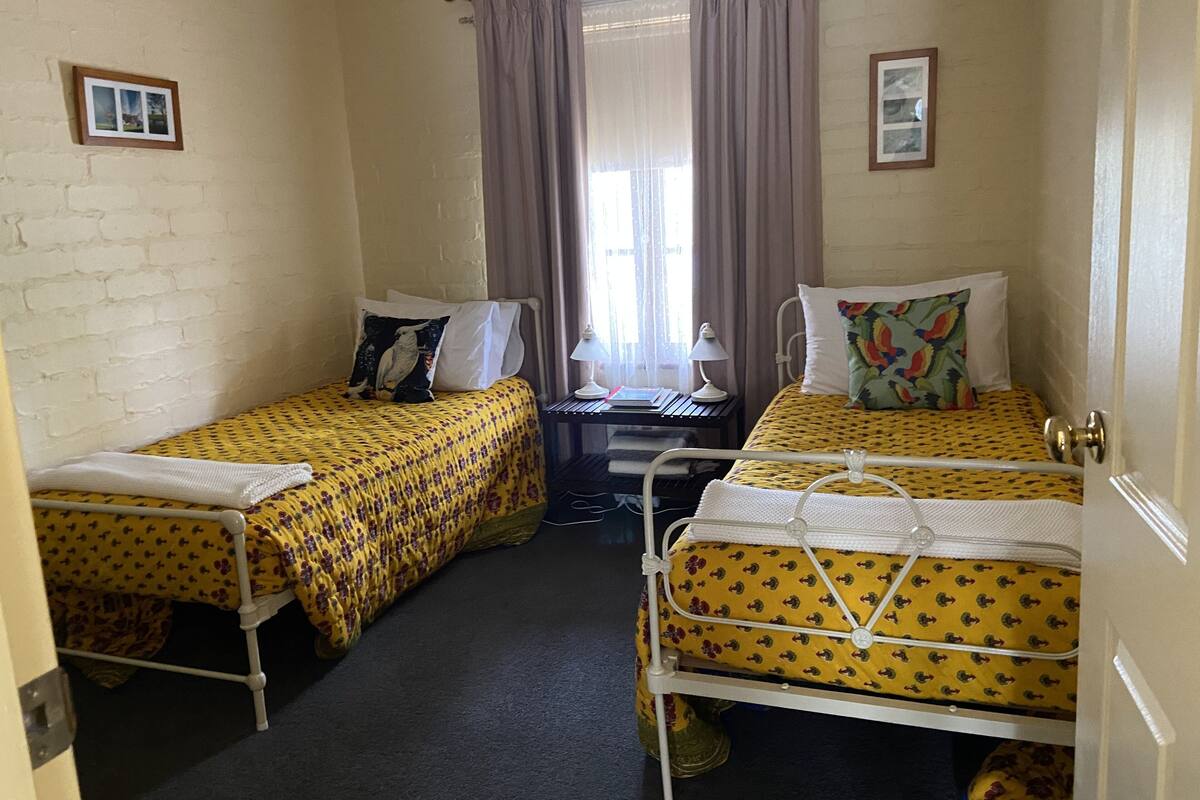 A small, compact room with two single beds, a desk with a chair, a window with curtains, and a door.