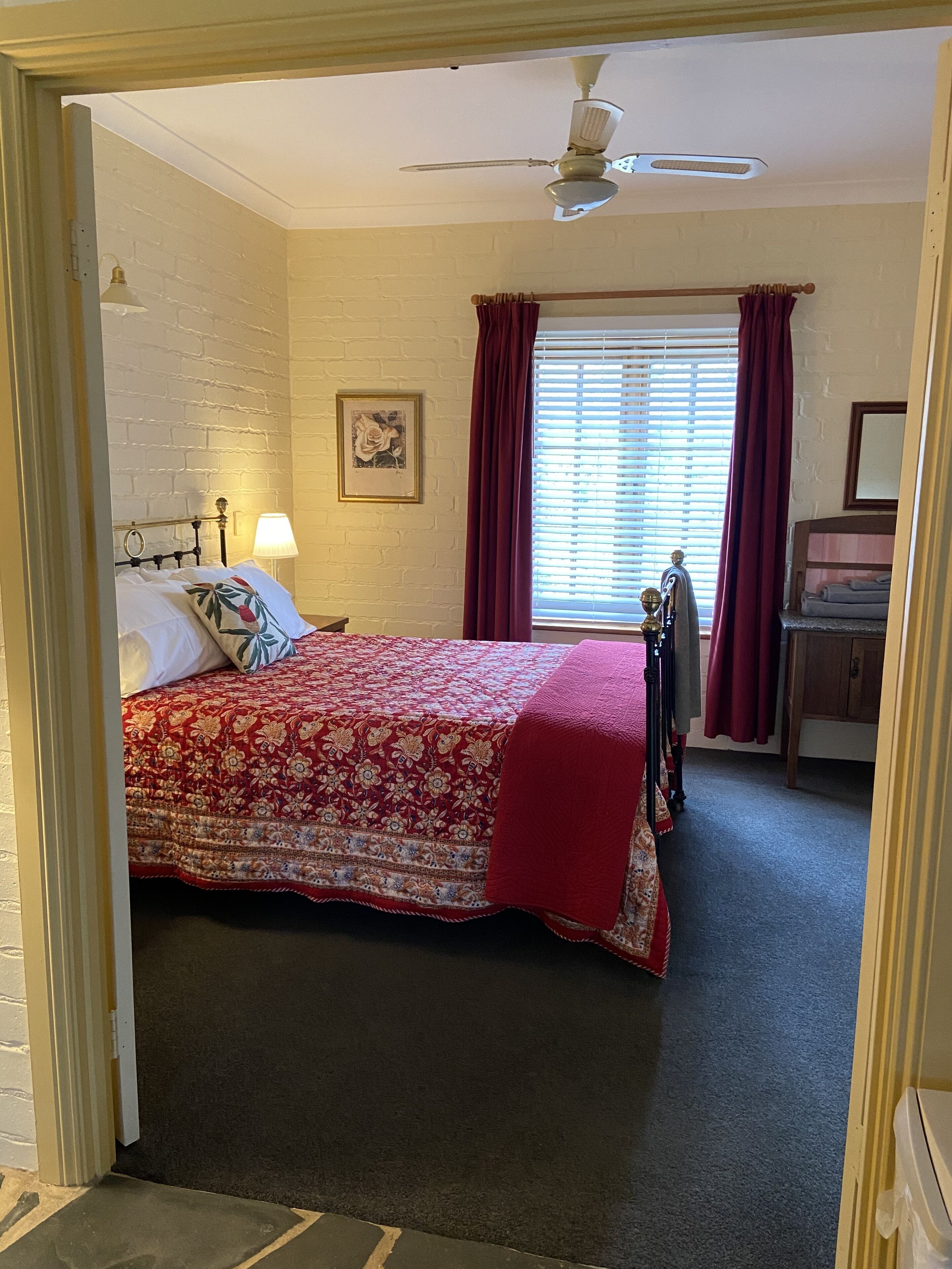 Een slaapkamer met een bed, nachtkastje, lamp, plafondventilator, raam met gordijnen en een kledingkast.